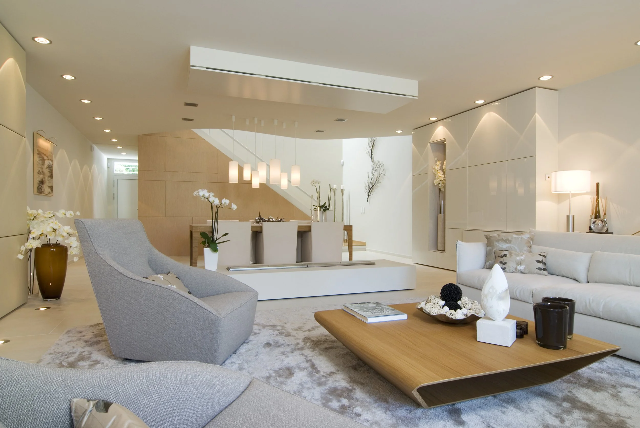 Moderne helle Wohnzimmer mit grauen und weißen Möbeln, einem großen weißen Sofa, einem niedrigen Holz-Couchtisch, mehreren weißen Orchideen, dekorativen Gegenständen und Lampen, im Hintergrund Treppe und Wanddeko.
