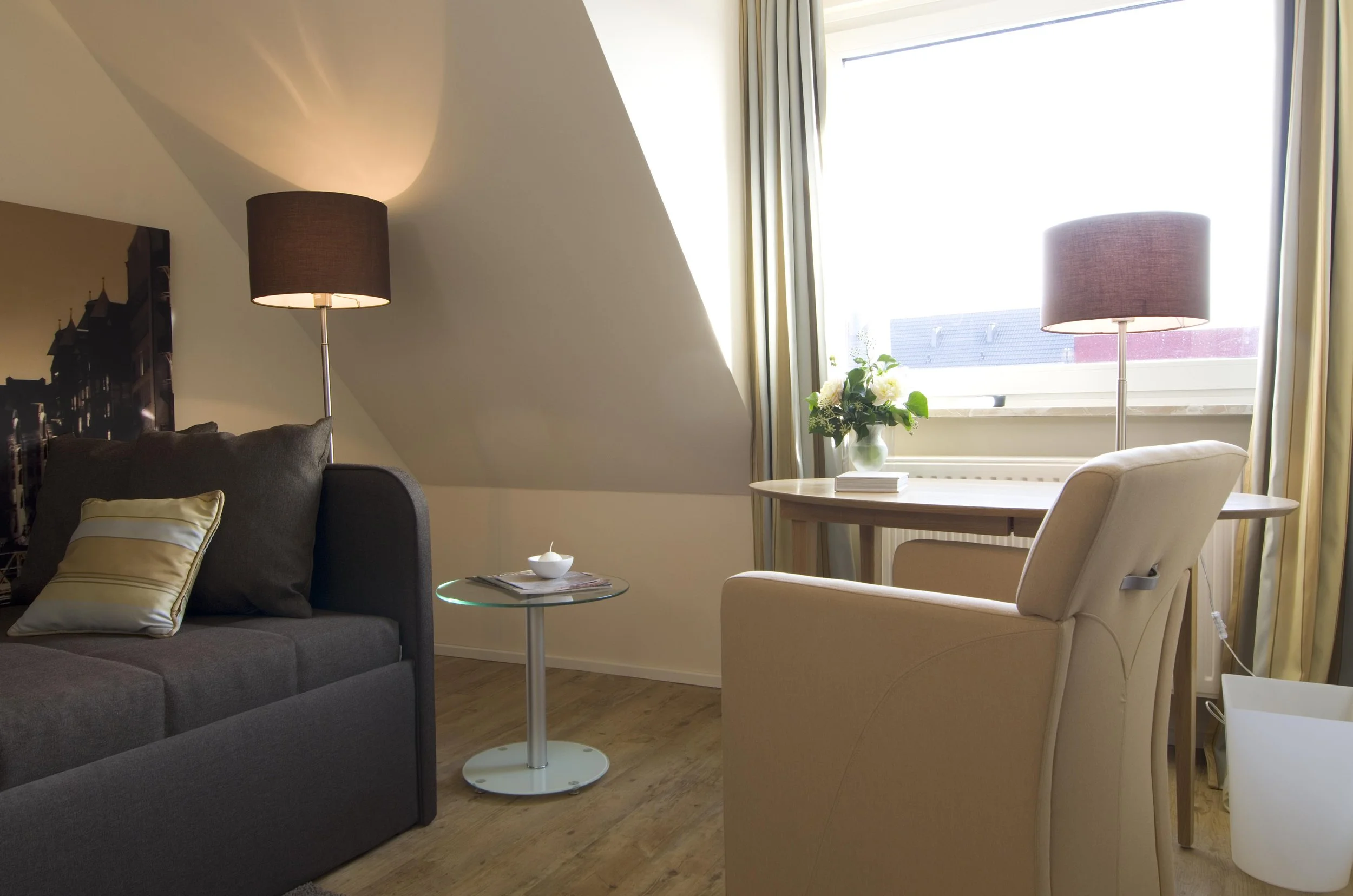 Gemütliches Wohnzimmer mit grauem Sofa, beige Armchair, Parkettboden, großen Fenstern mit Vorhängen, einem Beistelltisch, einem runden Esstisch mit Blumen, Lampen, und Deko