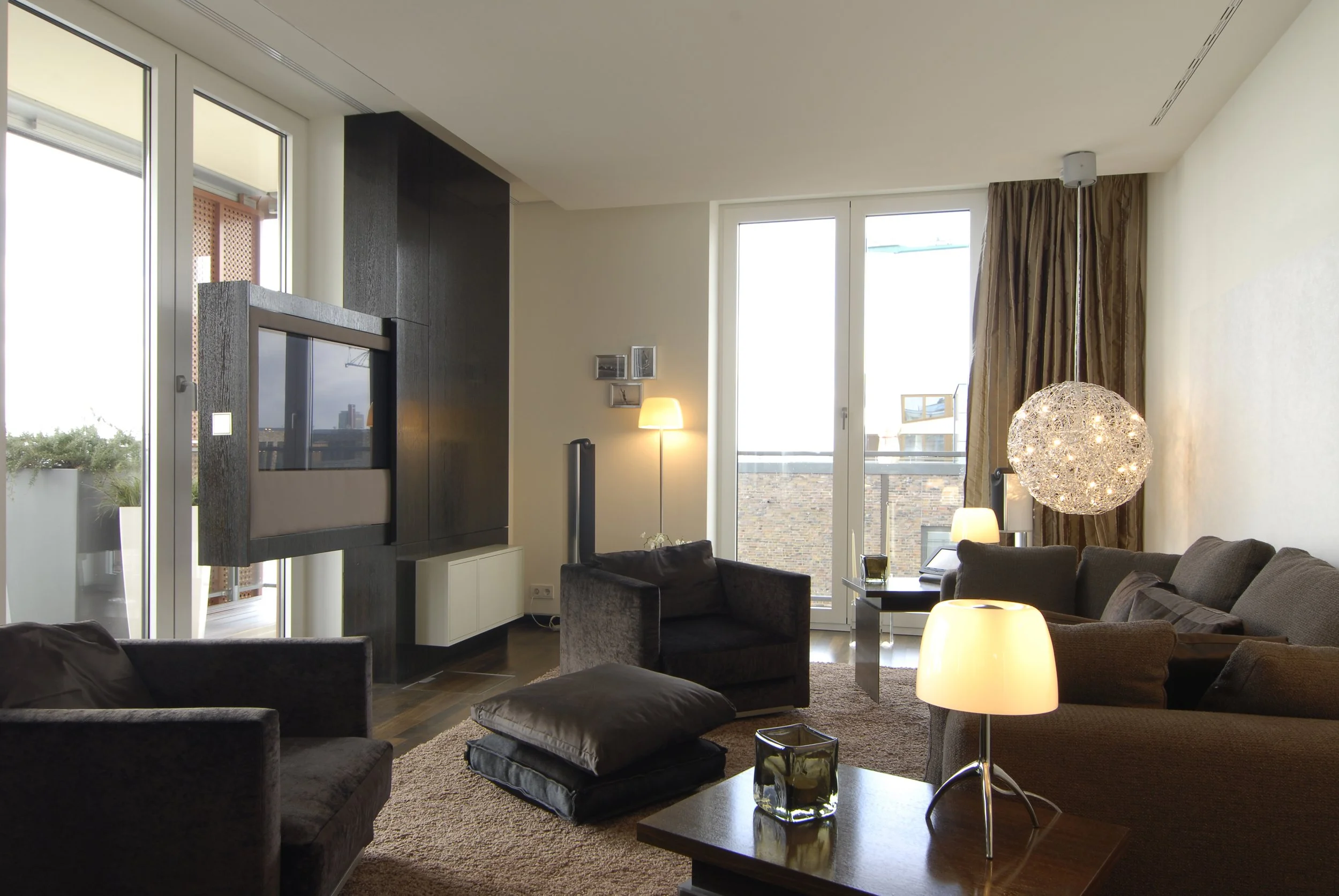 Gemütliches Wohnzimmer mit Sofa, Sessel, Couchtisch, Stehlampen, einem modernen Kristall-Lampe, Balkon, große Fenster und dunkler Wandpanel.