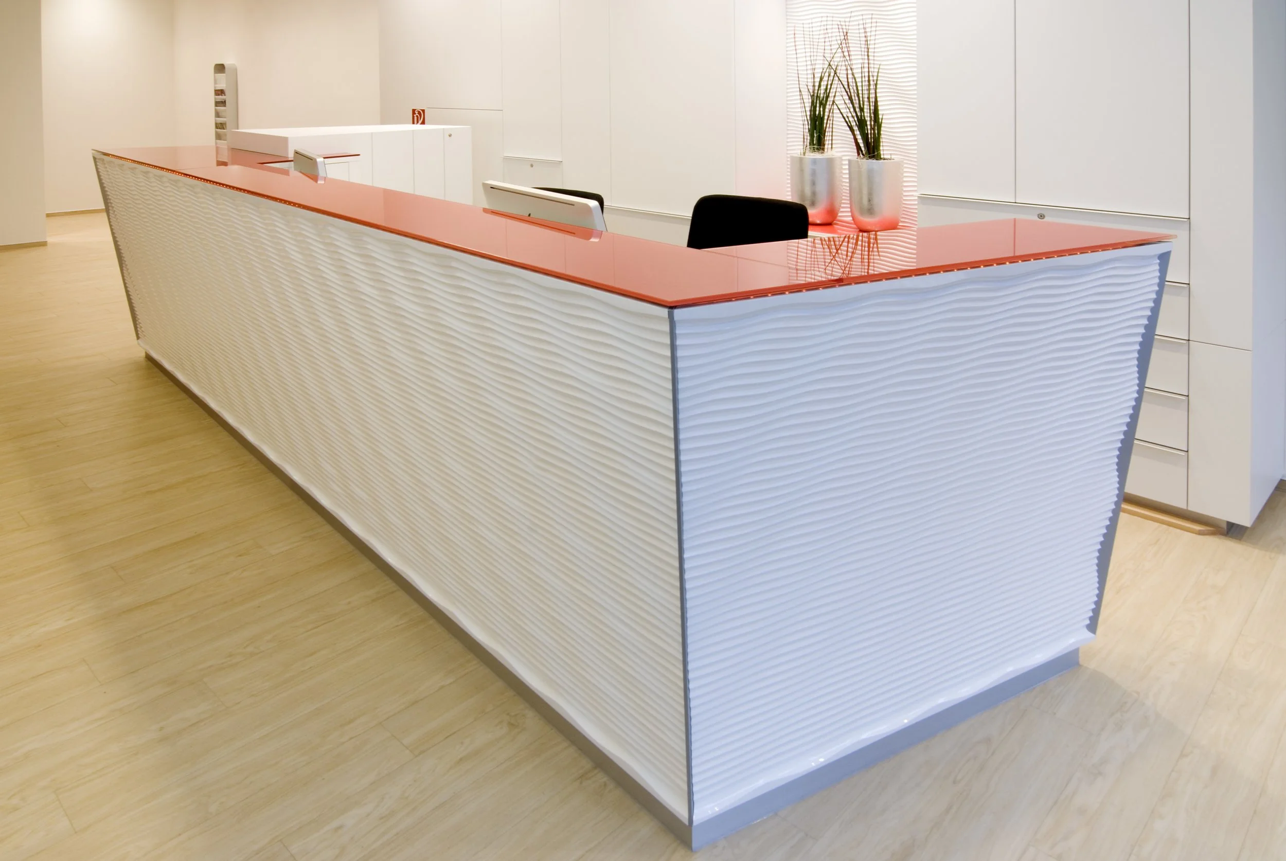 Moderne Kaffeebar mit weißen, gewellten Fronten und roter Oberfläche, zwei Pflanzen in silbernen Töpfen, im Hintergrund weiße Wände und Holzfußboden.