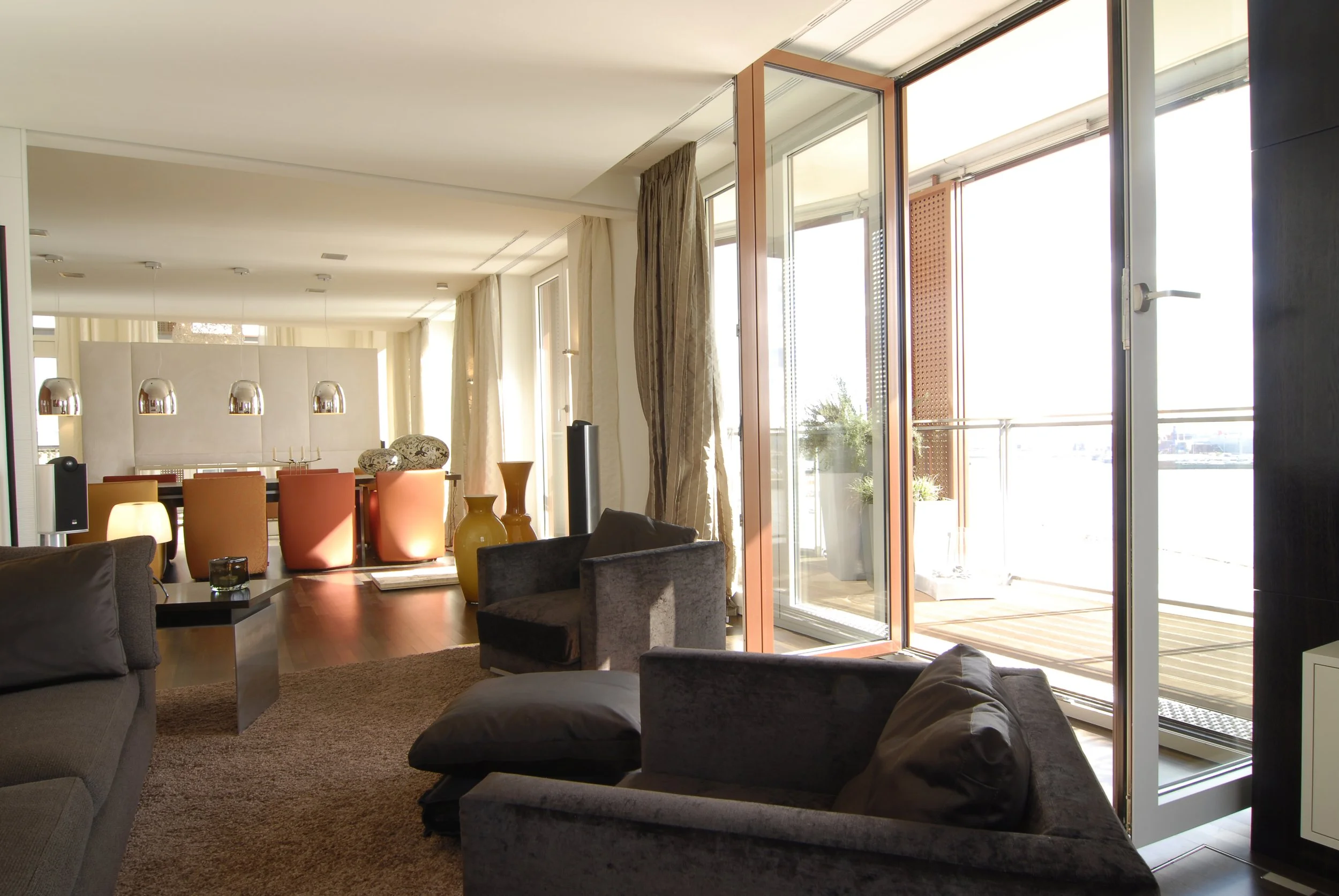 Helles Wohnzimmer mit großen Fenstern und Terrassentüren, gemütlichen Sofas, einem Teppich, Vasen und einem Essbereich im Hintergrund.