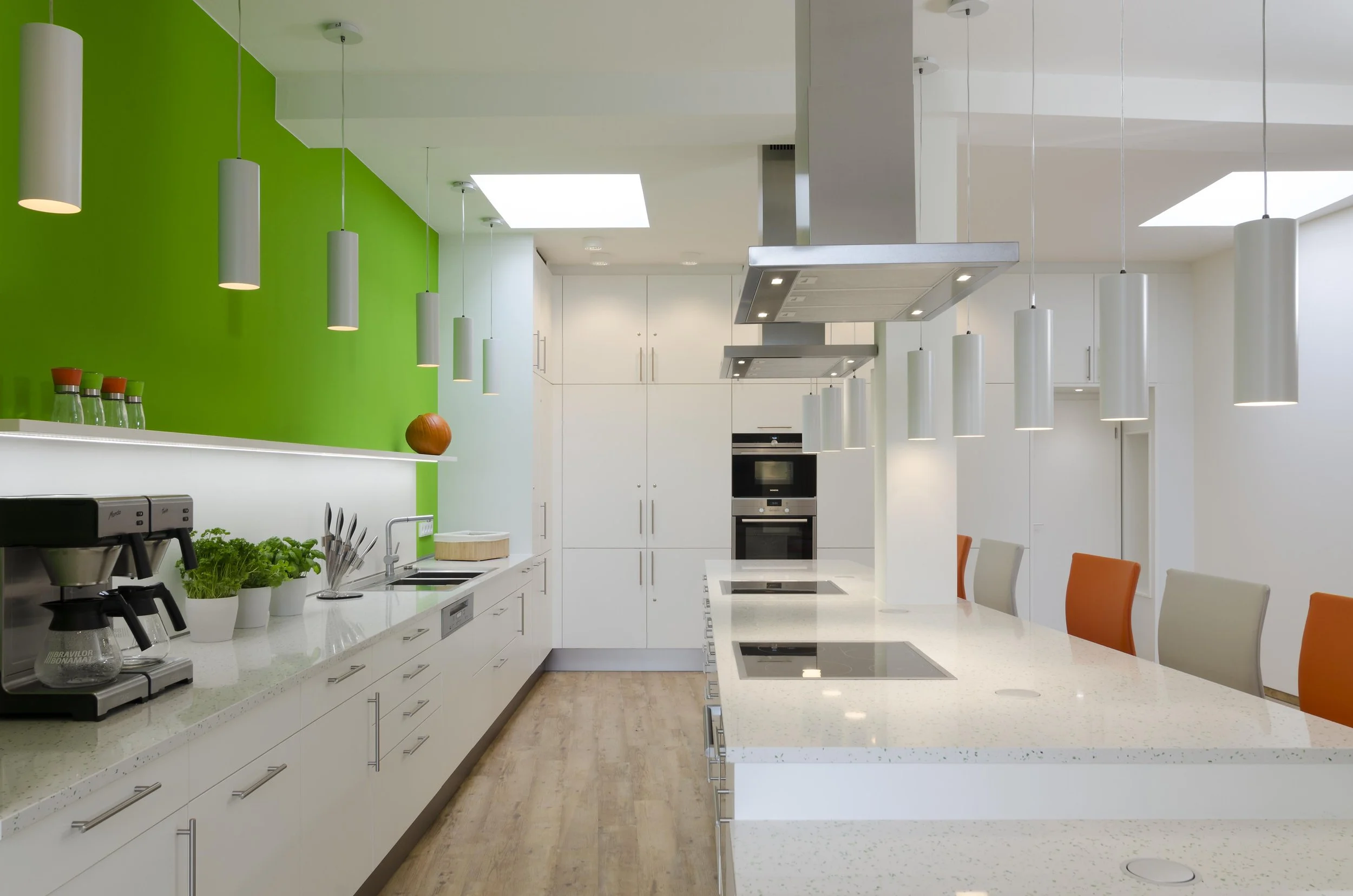 Moderne Küche mit weißen Schränken, grüner Wand, mehreren weißen Hängeleuchten, Küchenutensilien, Pflanzen und einem Frühstückstresen mit bunten Stühlen