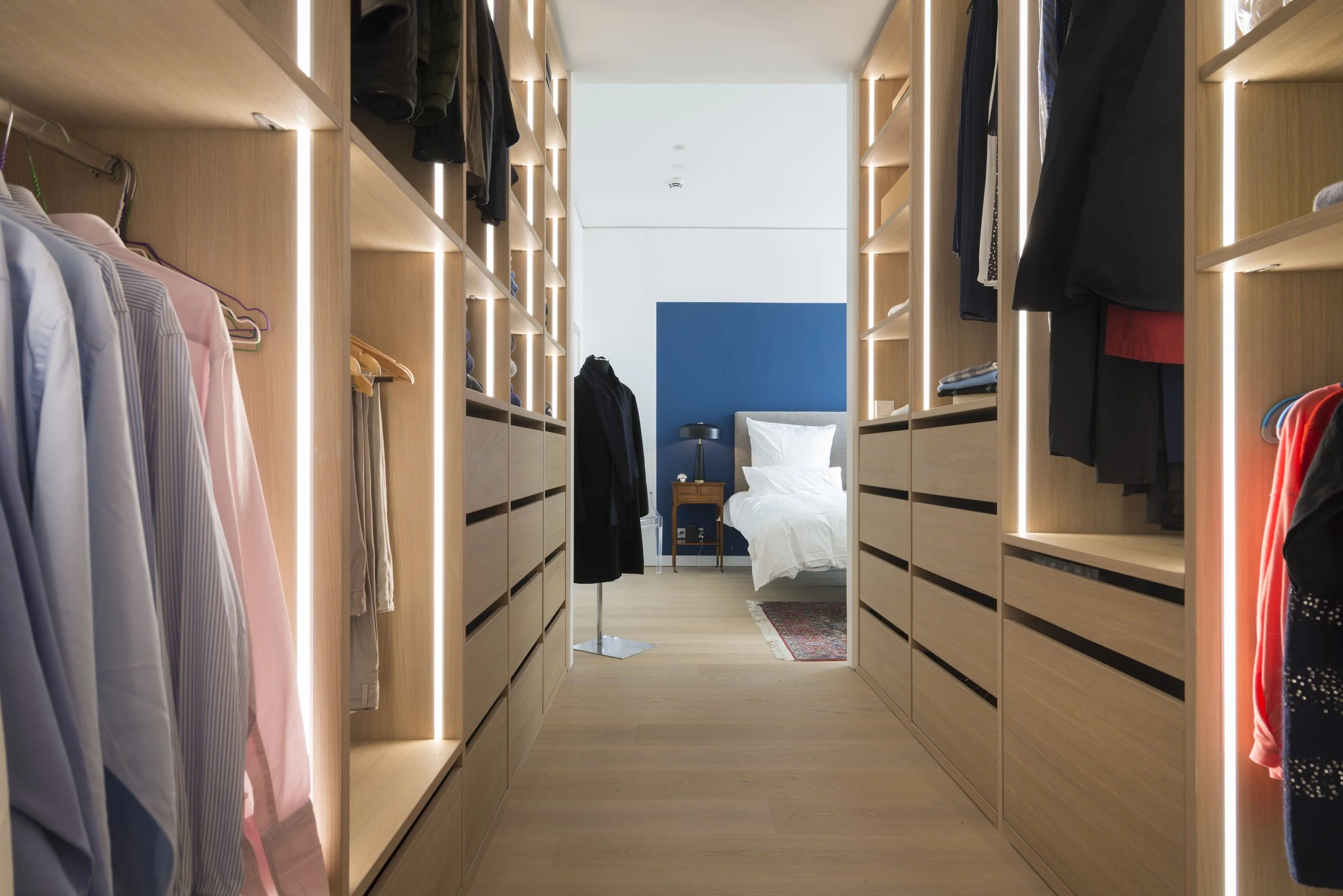 Innenaufnahme eines modernen begehbaren Kleiderschranks mit Holzregalen und Kleiderstangen, im Hintergrund ein Schlafzimmer mit Bett, Nachttisch, Lampe und blauer Wand.