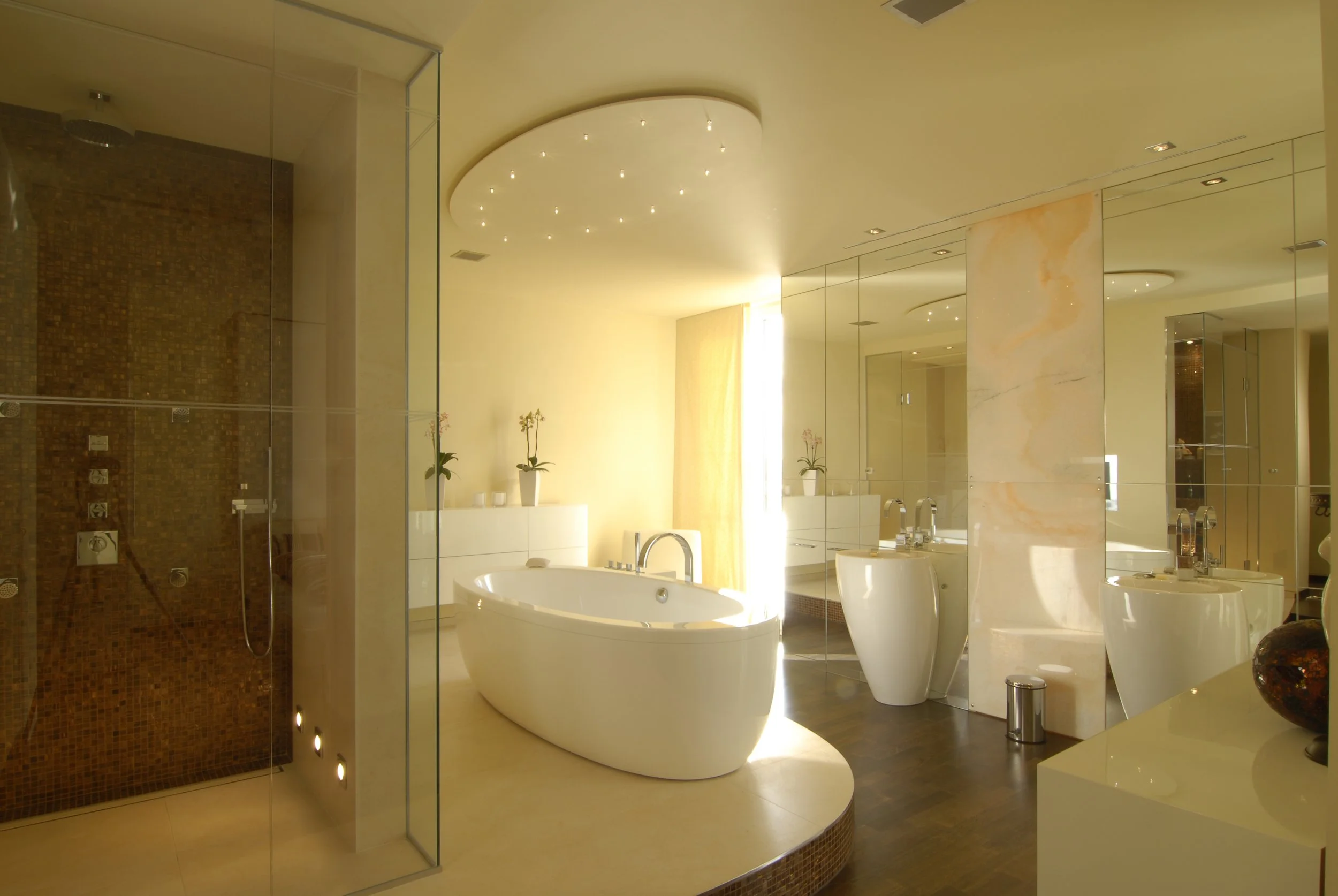 Luxuriöses Badezimmer mit freistehender Badewanne, separater Dusche, Badewannenarmaturen, Spiegelwänden, Pflanzen und indirekter Beleuchtung.