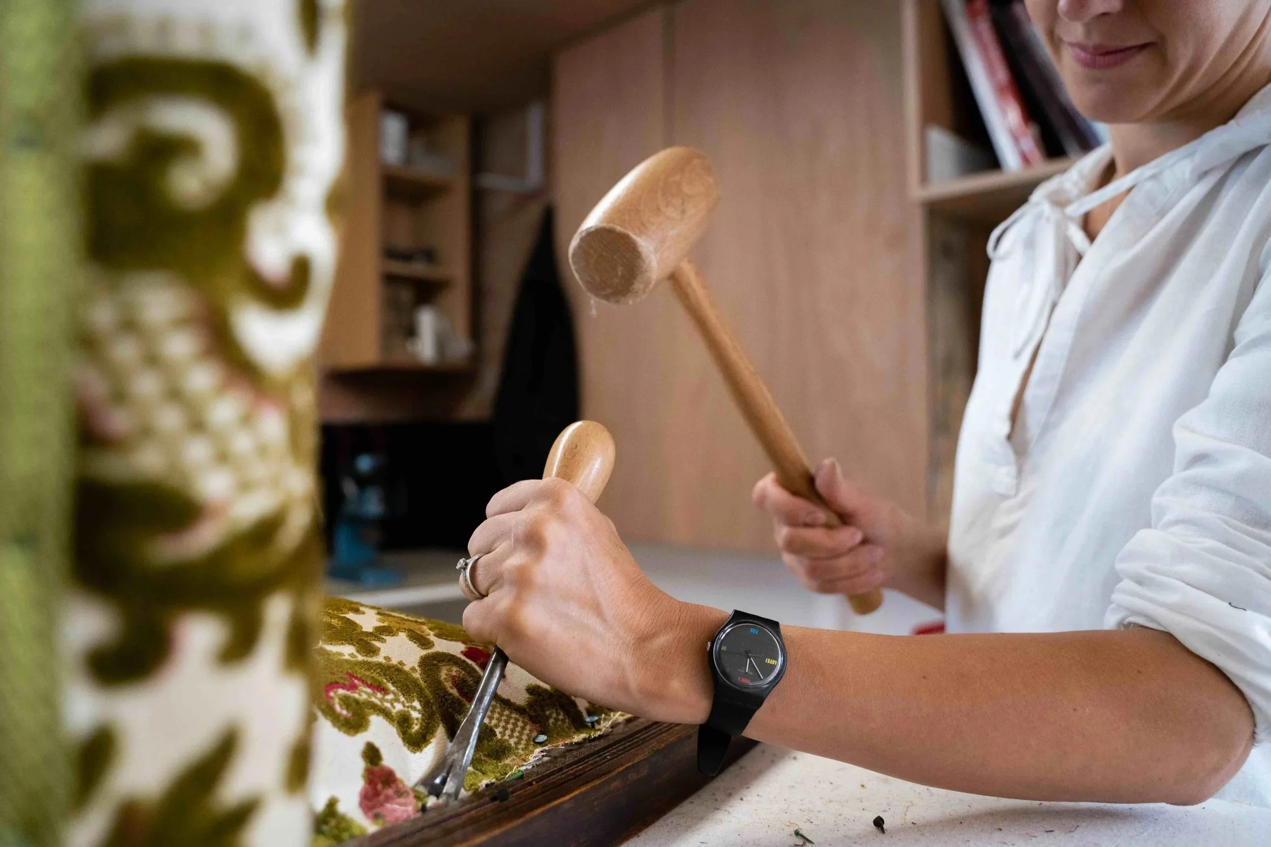 A person working on a textile project, using a hammer and chisel to manipulate fabric or embroidery on a wooden frame, with a patterned fabric on the workspace, wearing a black wristwatch.