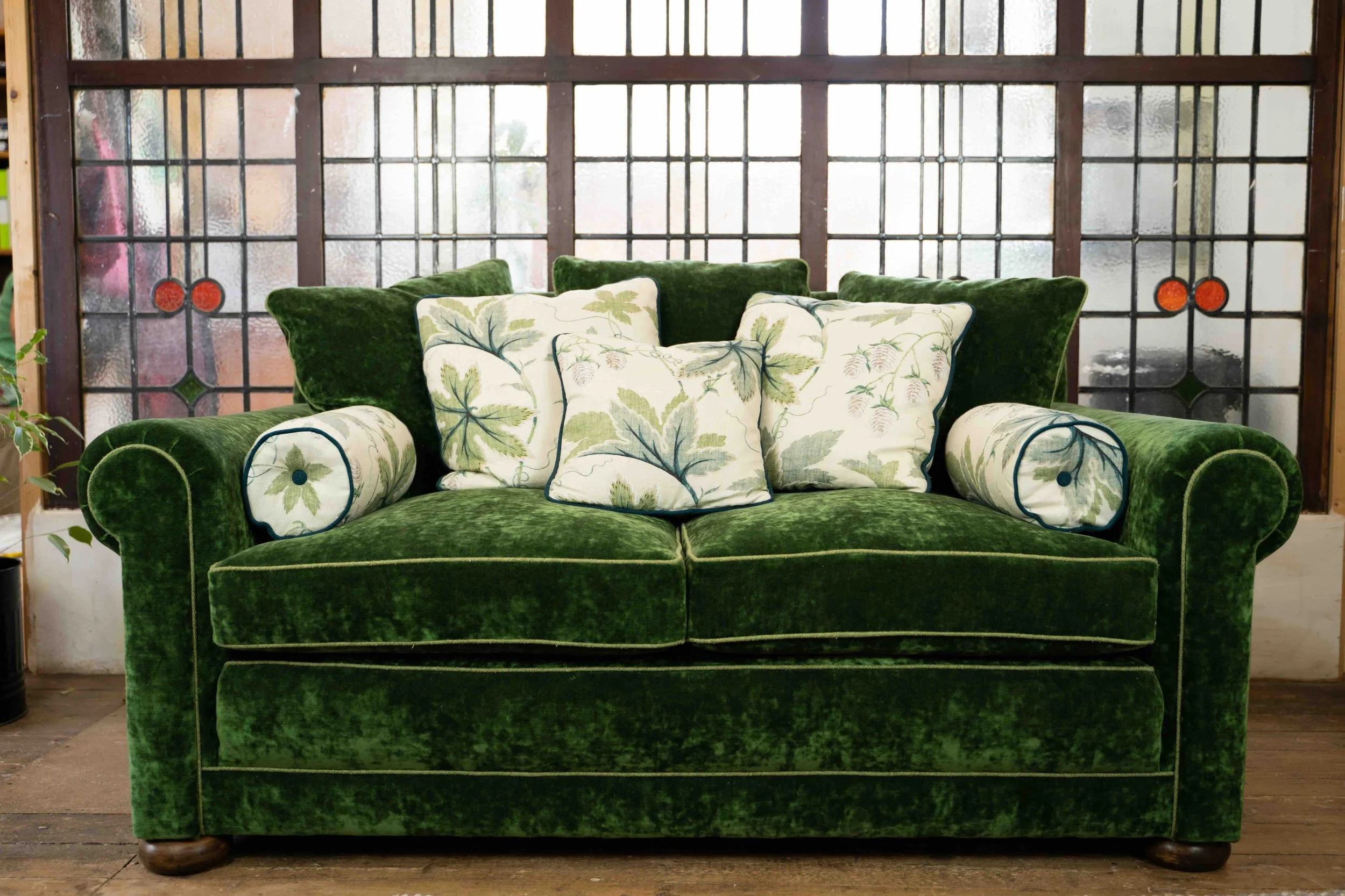 A green velvet sofa with decorative pillows, set against a window with a stained-glass pattern.