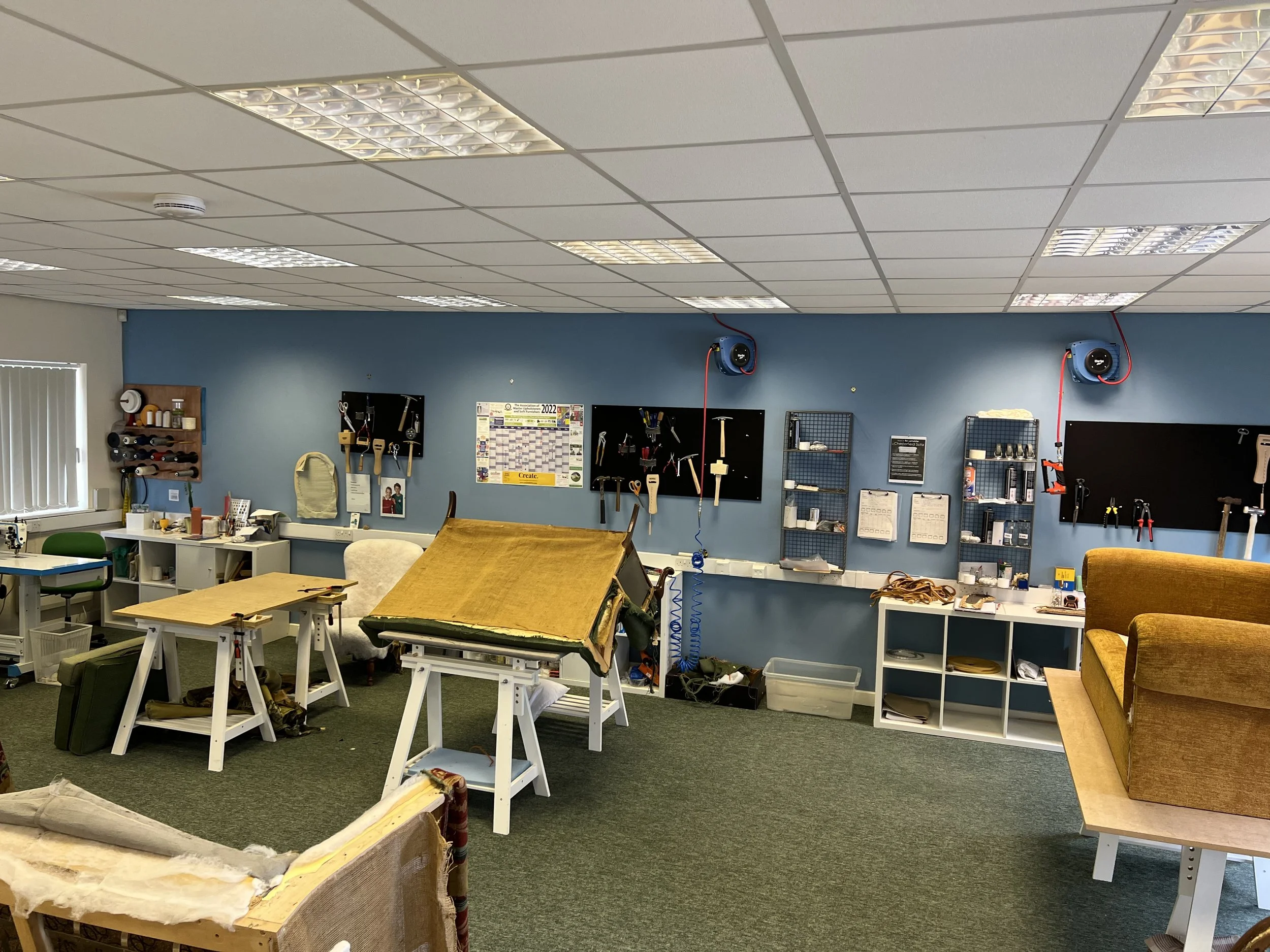 Workshop or craft room with workbenches, tools, and equipment, including a yellow upholstered chair and a workspace with tools and supplies on shelves.