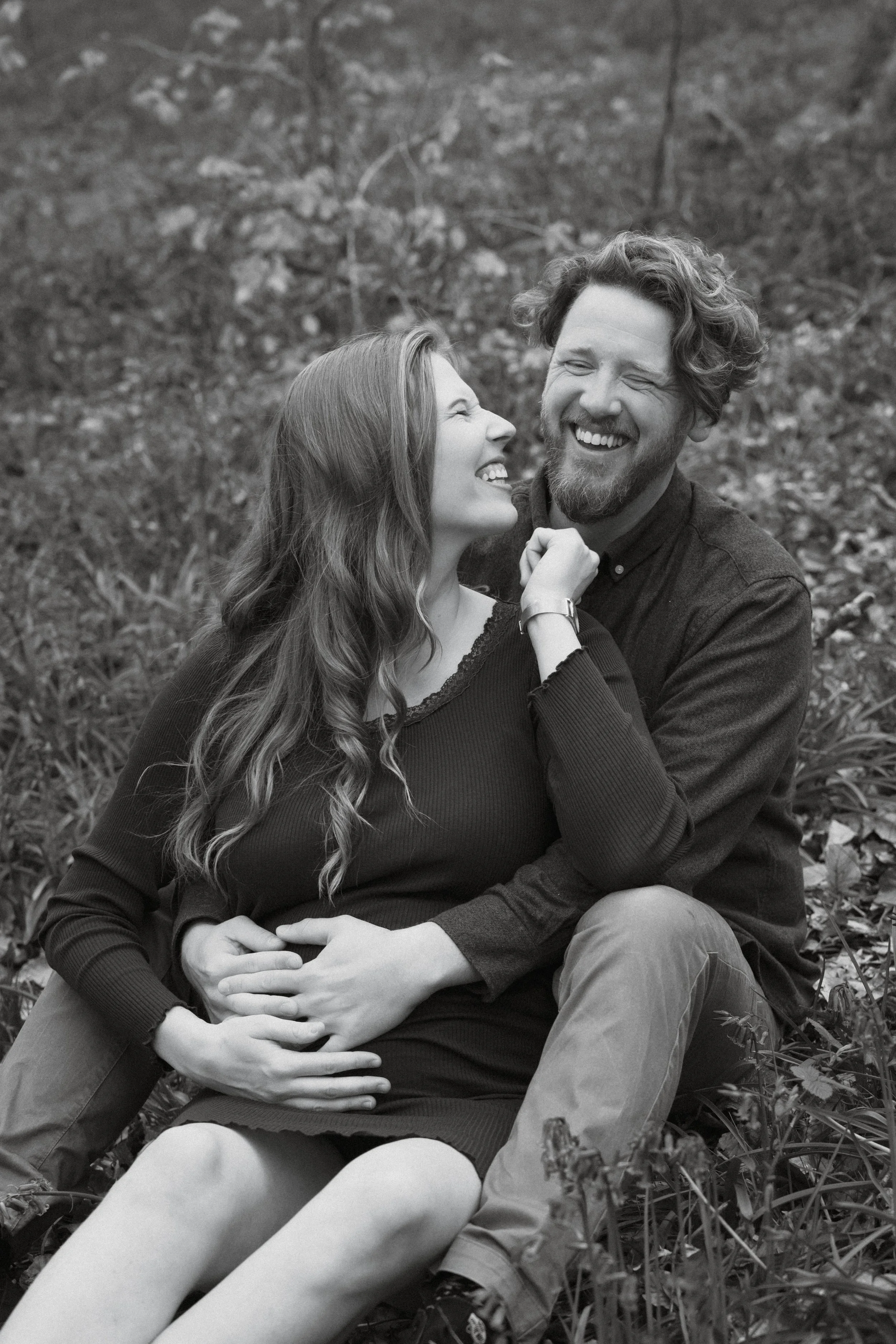 happy couple holding the woman's pregnant belly and laughing, sitting in nature during a coupe photoshoot