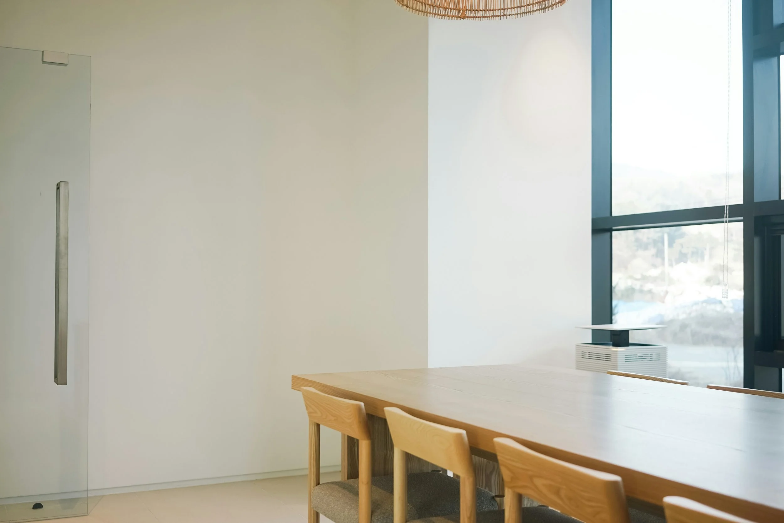 Interior de una sala de reuniones con una mesa de madera y sillas de madera y tela, ventana grande con vista al exterior y un ventilador de suelo.