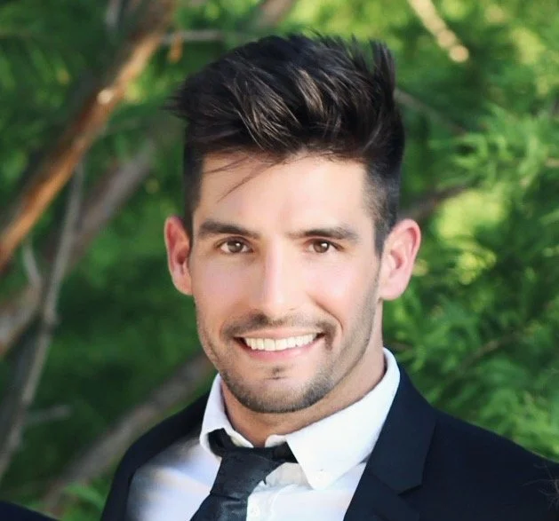 Man in a suit smiling with blurred green foliage background