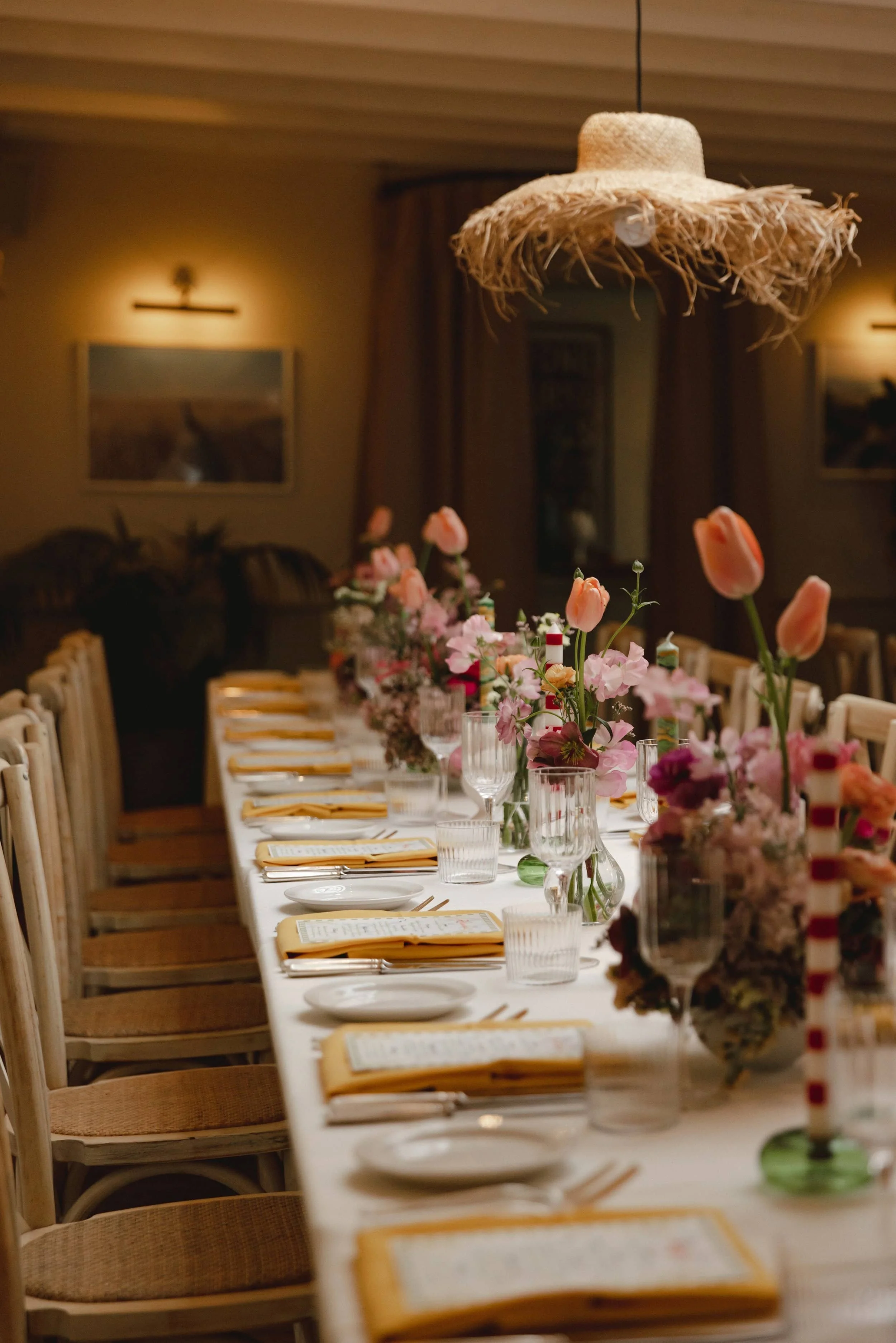 A long dining table decorated with pink and peach flowers in vases, with place settings including cutlery, glasses, and napkins, in a warmly lit room with framed artwork on the walls.
