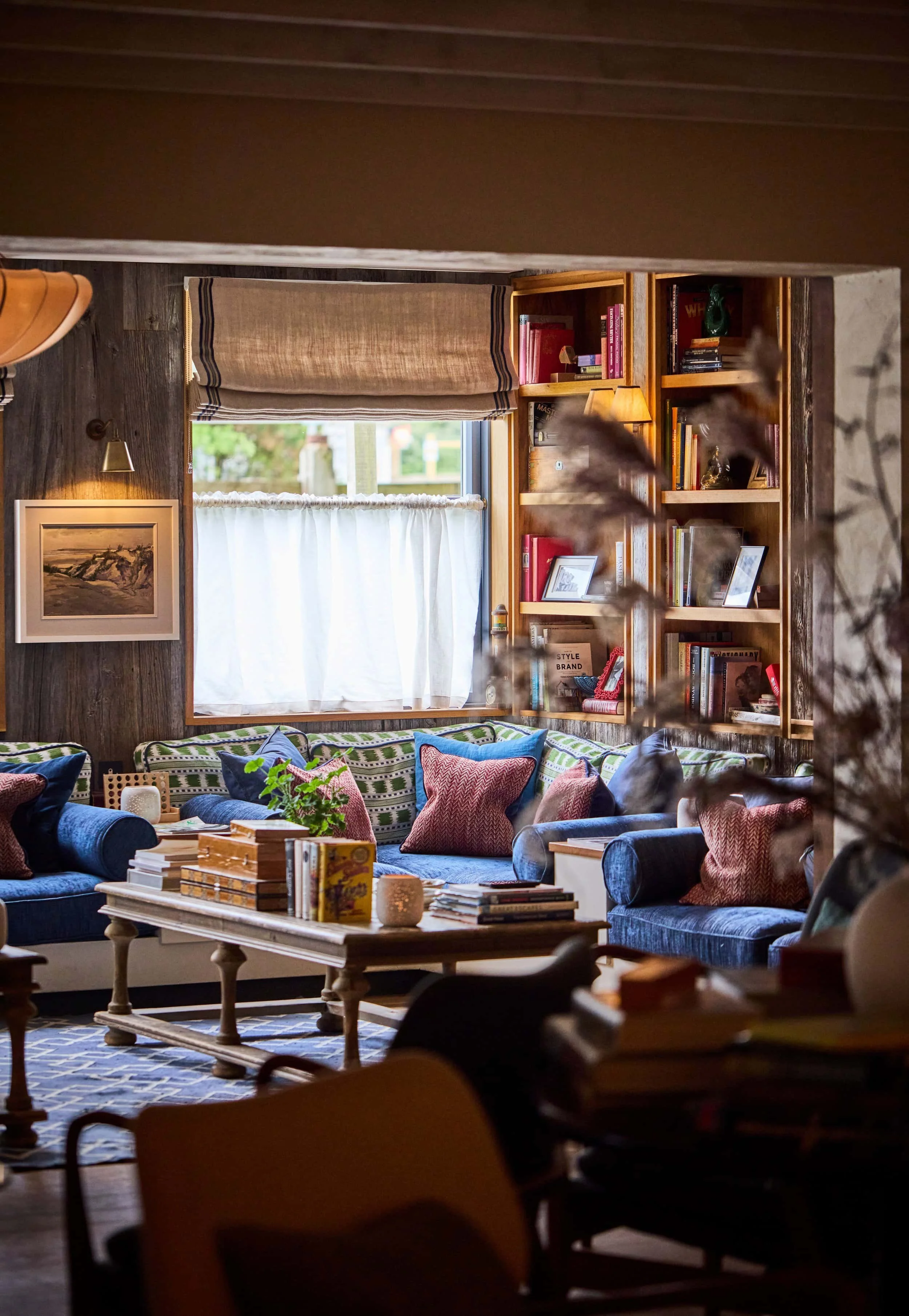 Cozy living room with colorful sofas, pillows, and coffee table filled with books and decorations. Window with shades, wooden shelves with books and decor, framed painting on wall, warm lighting.