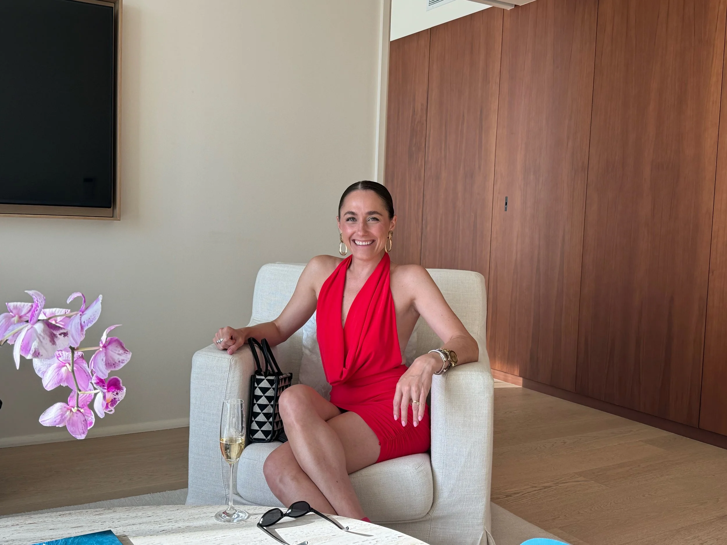 A woman in a red dress sitting on a white armchair, smiling, with a glass of champagne, sunglasses, and a black-and-white patterned bag on her lap, in a modern indoor setting.