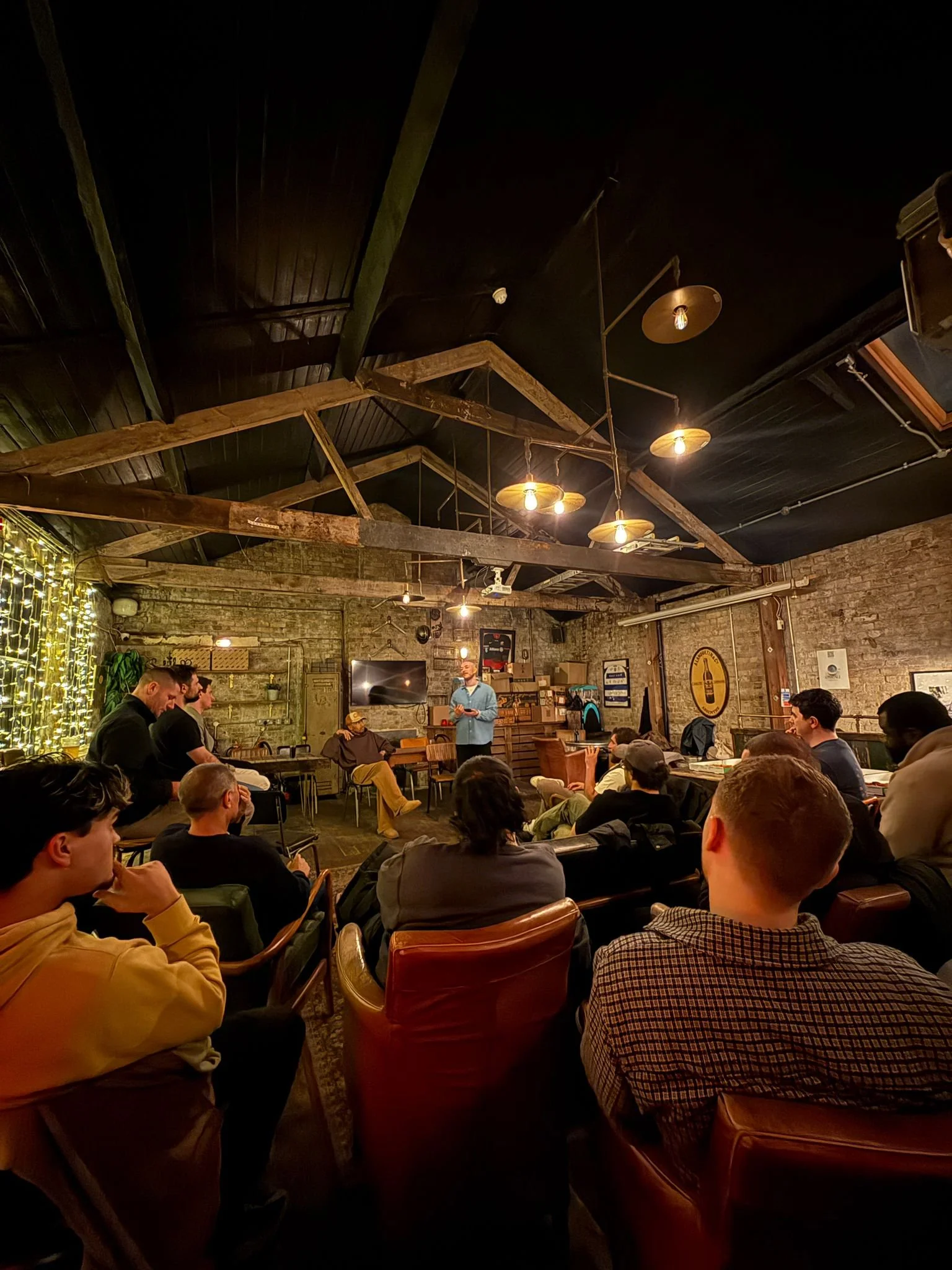 People sitting in a dimly lit restaurant or bar at the Wolf Pack london, listening to a speaker at a Good Chat event standing in front of a rustic brick wall, with exposed wooden beams and hanging industrial-style lights.