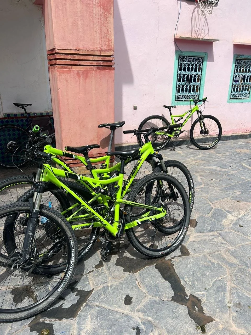 Drie groene mountainbikes geparkeerd op een stenen vloer voor een roze muur met twee ramen en een afbakeningskader in een tuin of patio.