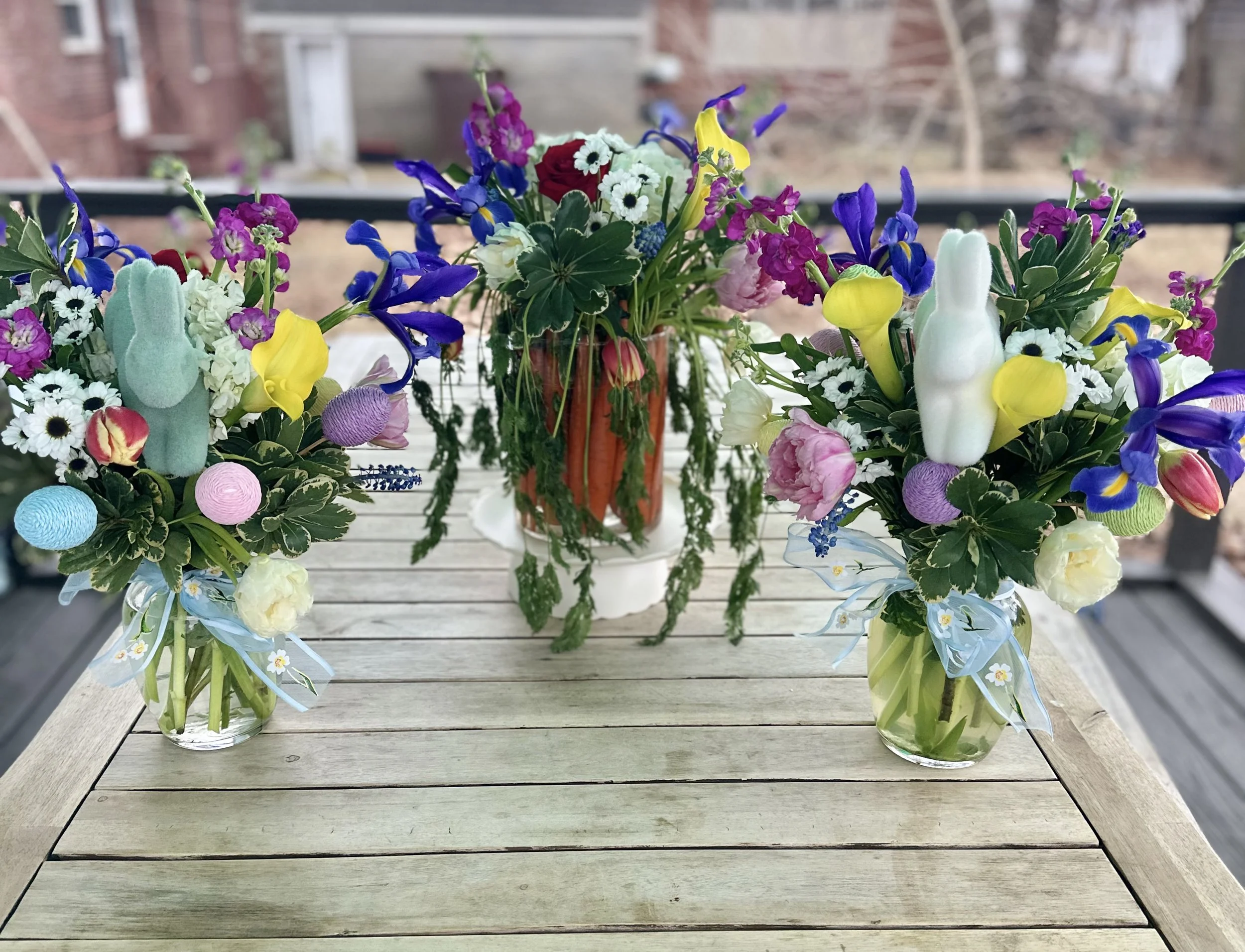 Spring floral arrangements with colorful flowers, decorated eggs, and bunny figures on a wooden table, featuring a central vase filled with carrots and greenery.