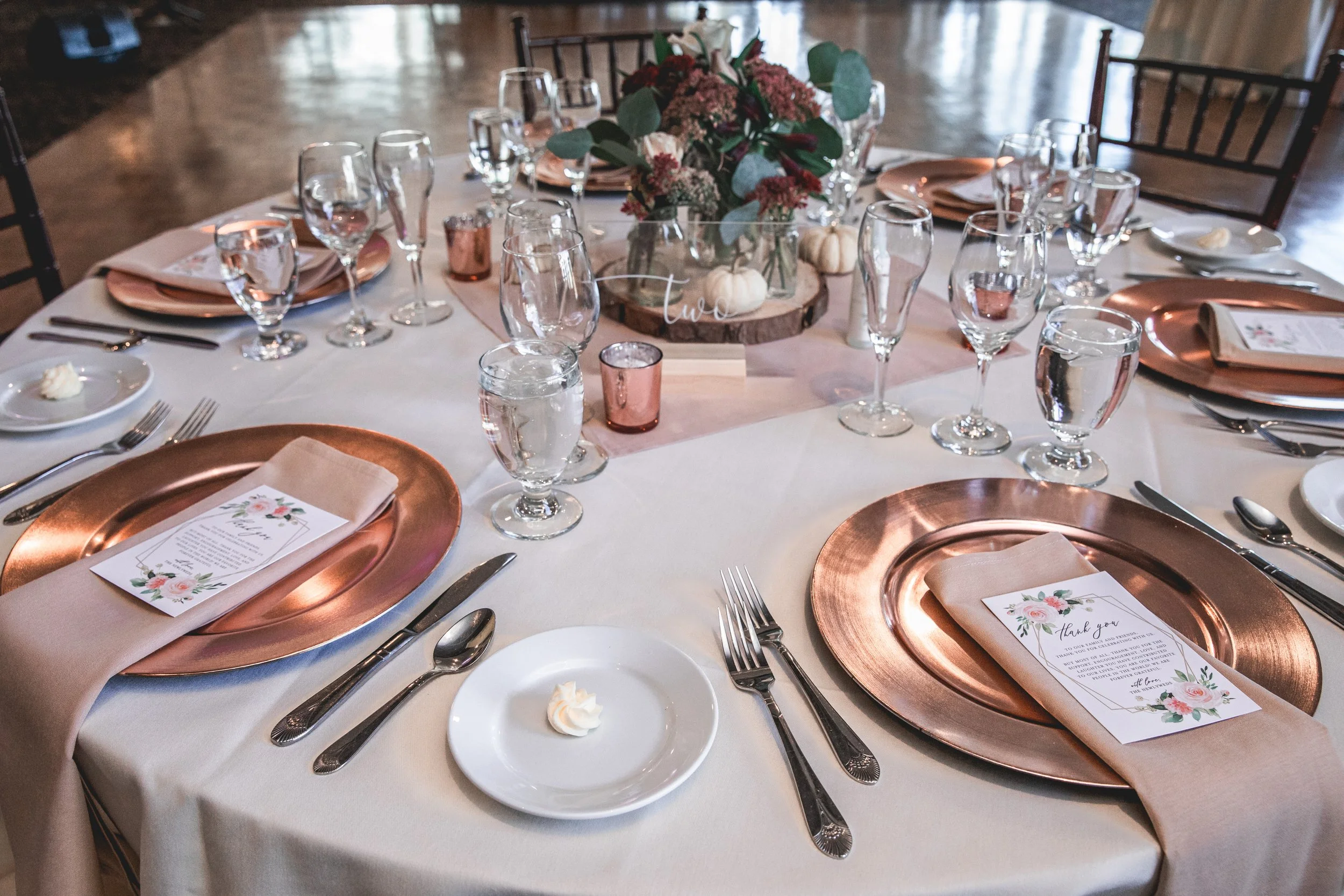 Elegant table setting with copper charger plates, white napkins, floral menus, glassware, and a floral centerpiece.