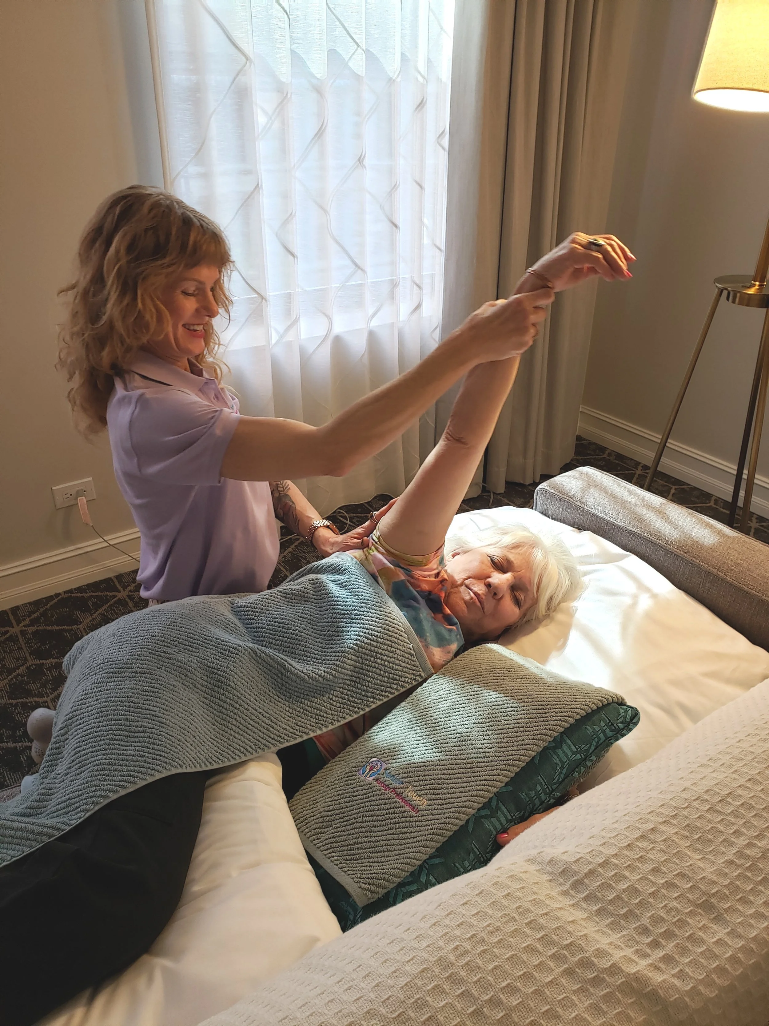 An elderly woman lying in bed, stretching her arm while a woman standing next to her helps assist. The elderly woman appears to be in a well-lit hotel room, with curtains and a bedside lamp visible.