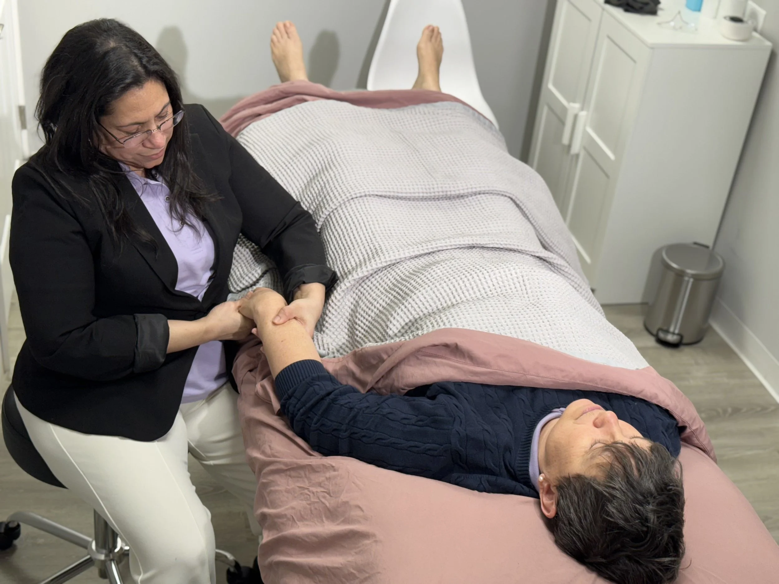 A woman in a blazer and glasses is holding the hand of a patient lying on a medical examination table, in a clinical setting.