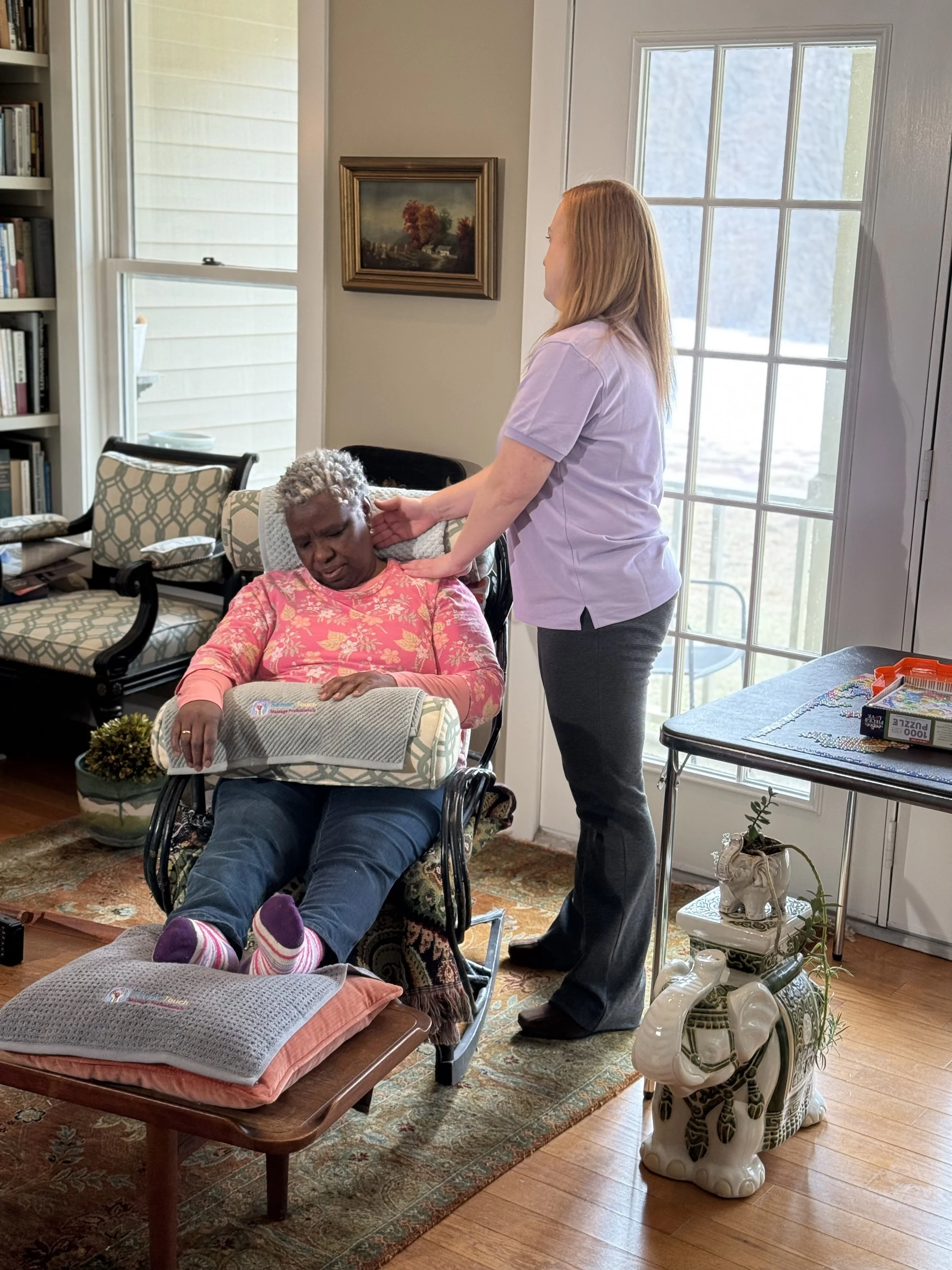 A woman with gray hair receives a massage on her neck from a caregiver in a living room. The woman is sitting in a reclined chair, covered with a pink floral shirt, and has her eyes closed. The caregiver, standing beside her, is wearing a purple shir