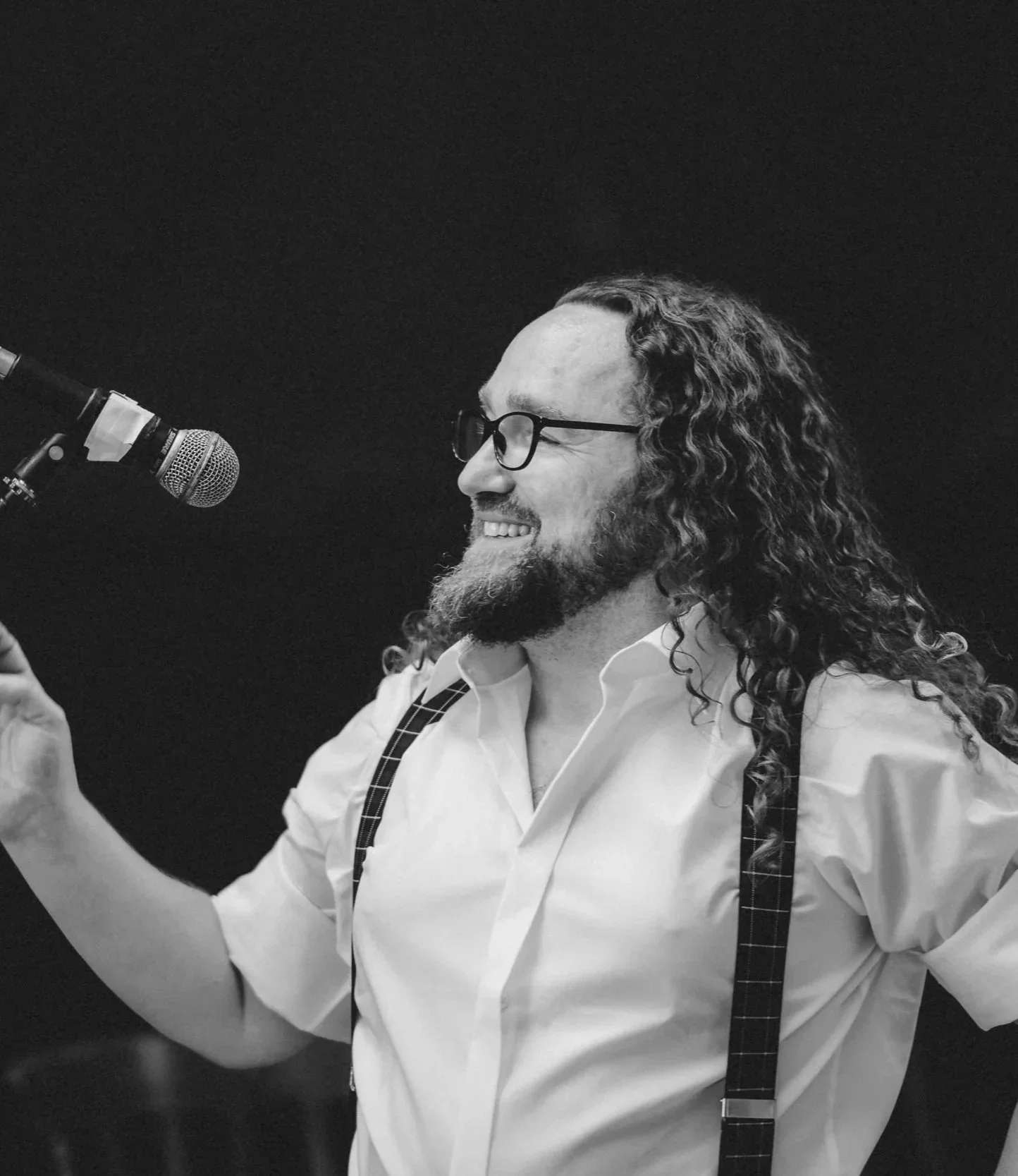 Black and white photo of John McGuirk-Wilding smiling  with long curly hair and glasses, wearing a white shirt with suspenders, speaking into a microphone.