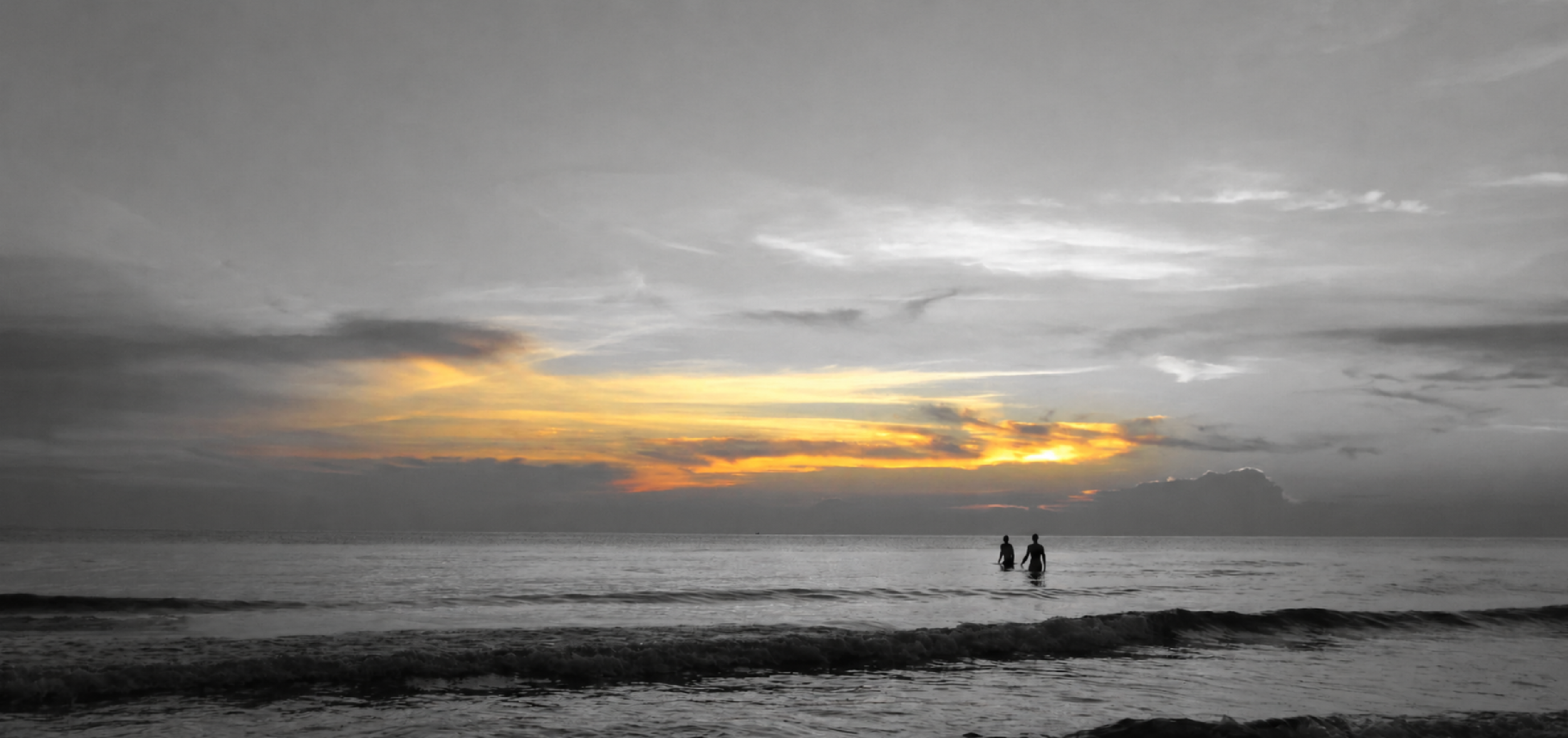 Two people standing in the ocean at sunset with a cloudy sky, some orange and yellow hues near the horizon, and waves at their feet.