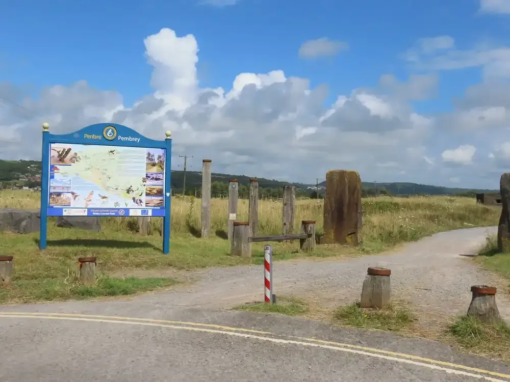 View of a park entrance with a large map sign, wooden barriers, and a gravel path leading into the park under a partly cloudy sky.