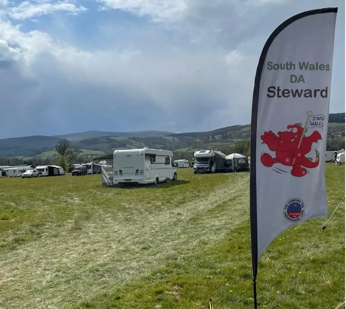 A campground with caravans and camper vans on a grassy field, with rolling hills and cloudy sky in the background. A flag with the words 'South Wales DA Steward' and a cartoon red dragon holding a South Wales flag is prominently displayed in the foreground.