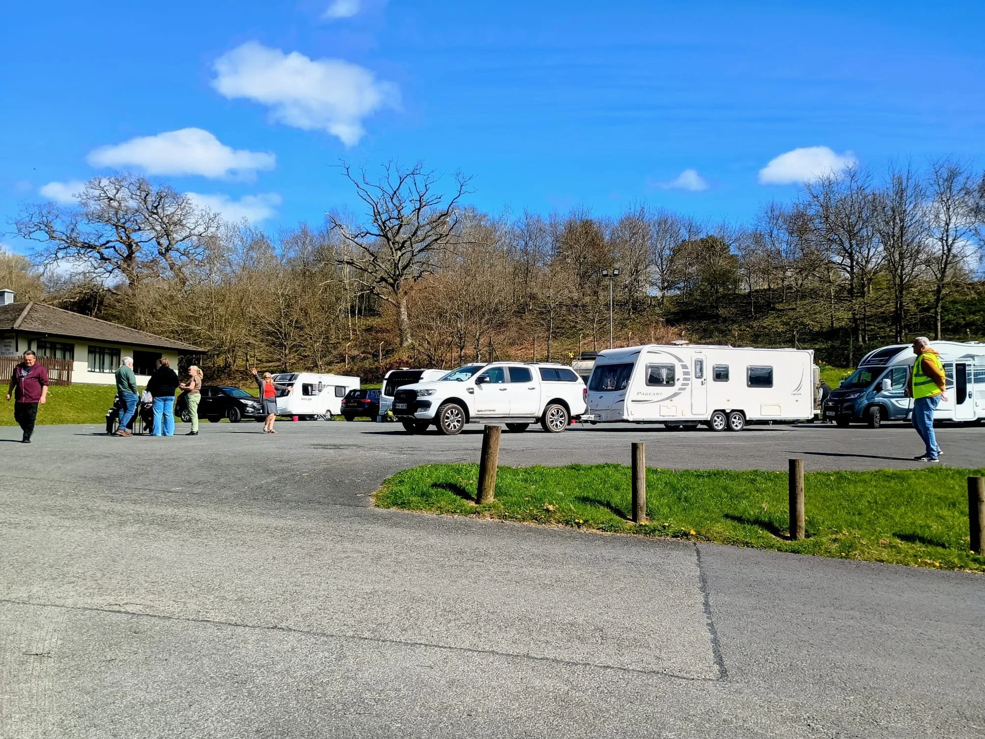 People gathering in a parking lot with several cars and camper vans under a blue sky with clouds and leafless trees in the background.