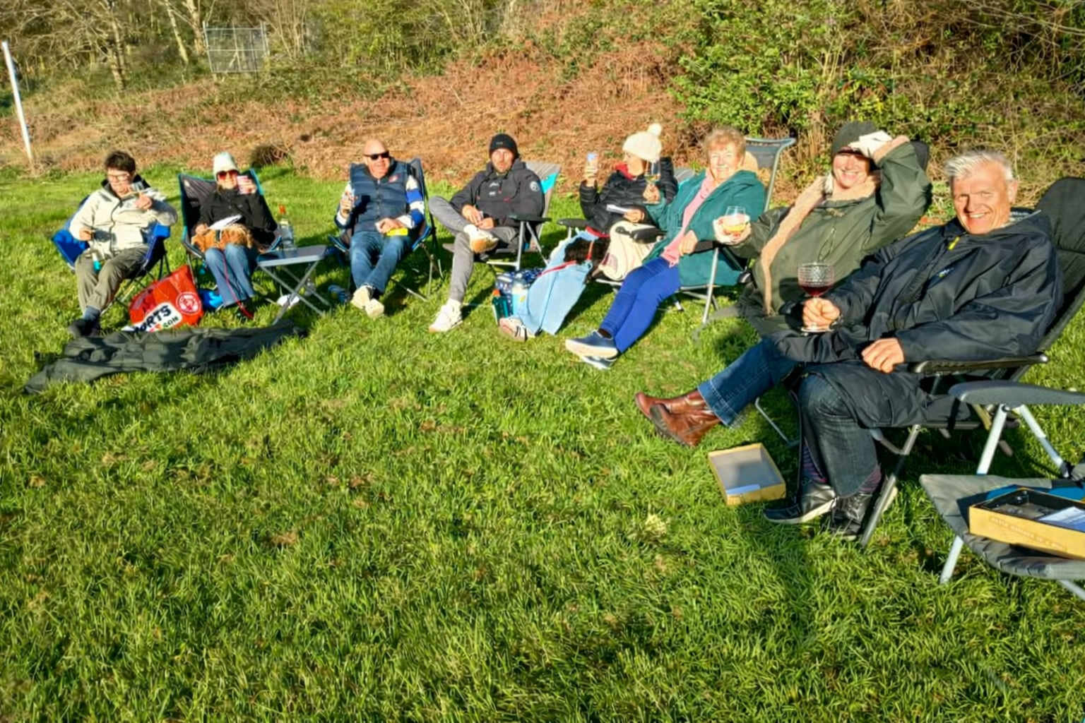 A group of nine people sitting on foldable chairs in a grassy outdoor area, enjoying drinks and each other's company on a sunny day.