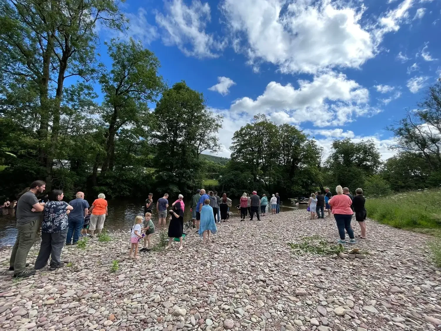 A group of people gathered along the rocky shore of a river, with trees and a partly cloudy sky in the background.