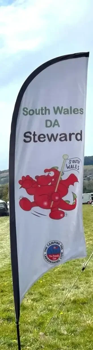 A tall, curved flag displays the text "South Wales DA Steward" with a cartoon red devil holding a flag that says "South Wales." The flag is set outdoors on a grassy area with a few parked cars in the background.