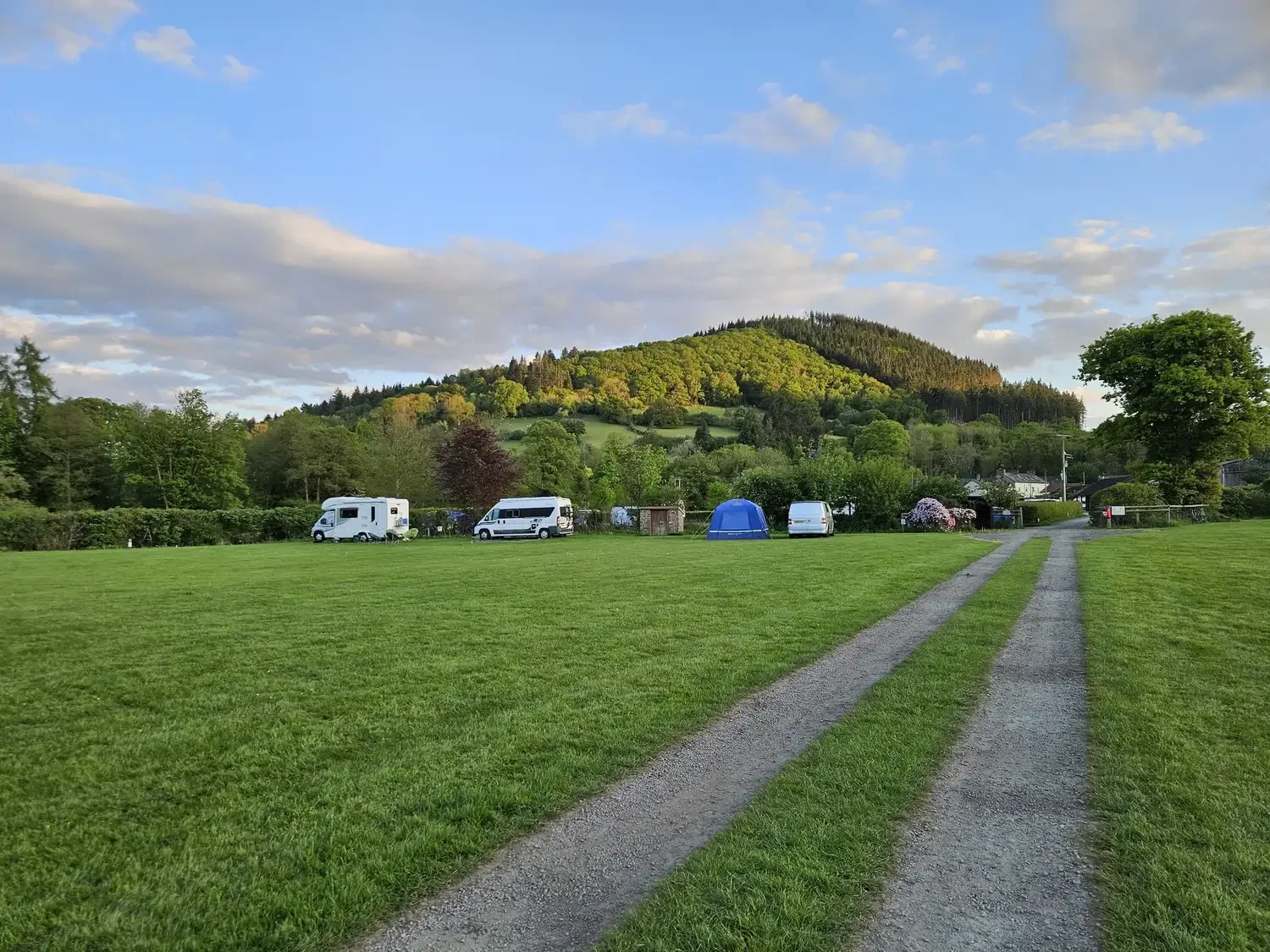 A scenic view of a campground with a dirt road leading to parked campers and tents, green grass, trees, and a hill in the background under a partly cloudy sky.