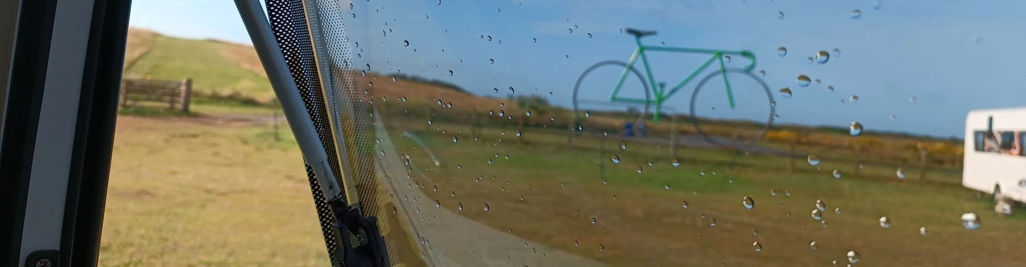 Rain droplets on a window with a distant green bicycle sculpture in the background, outdoor grassy area, and cloudy sky.