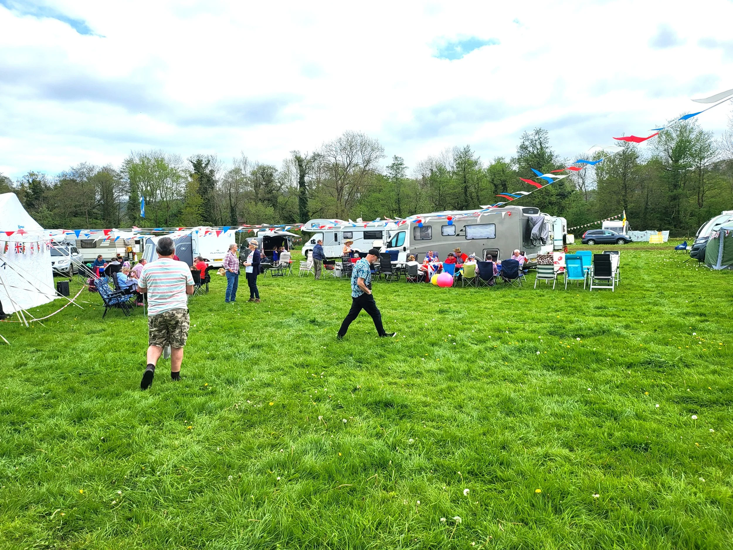 A lively outdoor gathering with people socializing and enjoying themselves among caravans and tents decorated with red, white, and blue bunting, set on a grassy field with trees in the background under a partly cloudy sky.