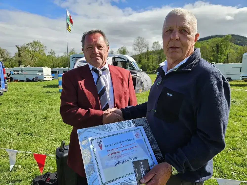 Two men shaking hands outdoors, one holding a certificate of recognition, at a camping or caravan site with tents, caravans, and a flag in the background.