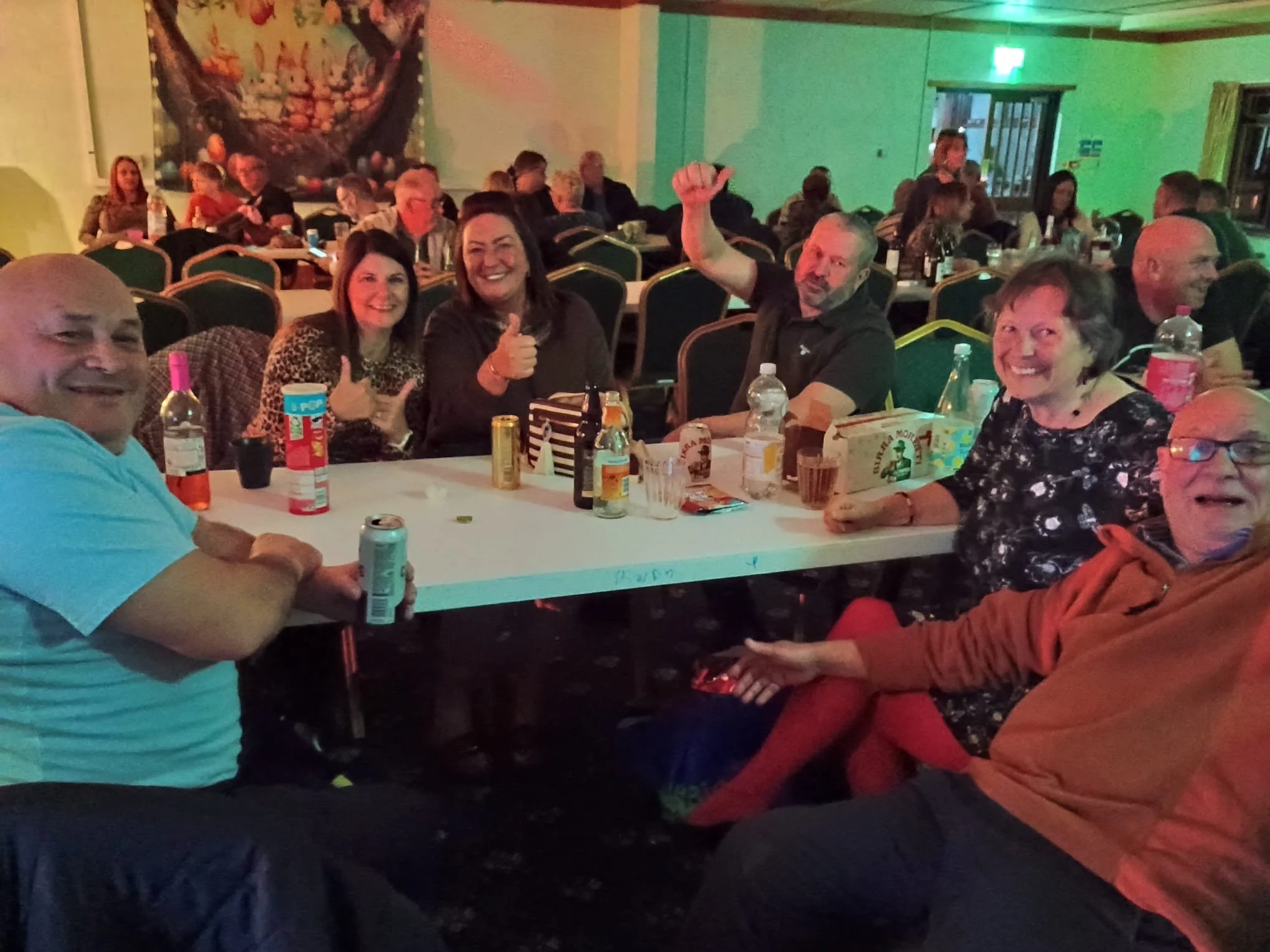Group of people sitting around a table at an indoor event, smiling and enjoying drinks, with some giving thumbs-up gestures.