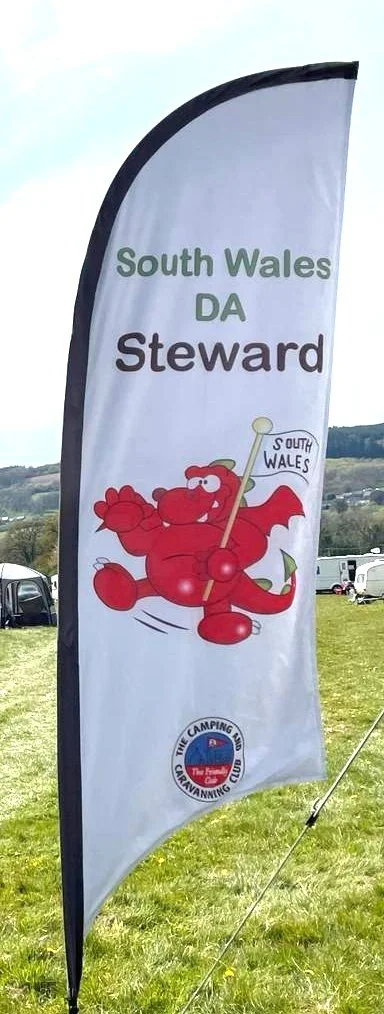A vertical flag banner displaying the South Wales DA Steward logo, featuring a red cartoon dragon holding a South Wales flag, set against a background of grass and parked vehicles.