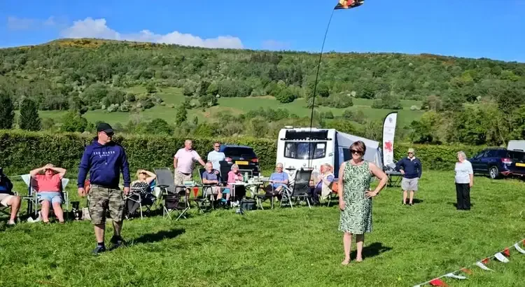 Group of people at outdoor gathering on a grassy field, with a camper, cars, and hills in the background, under a partly cloudy sky.