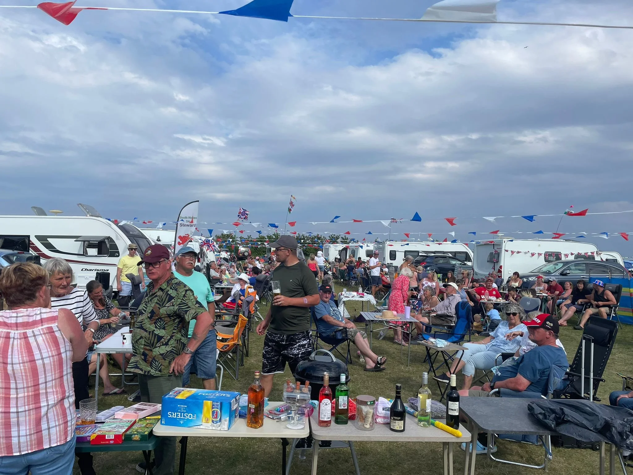 Crowd of people enjoying an outdoor event with camping trailers and Union Jack flags, under a partly cloudy sky.