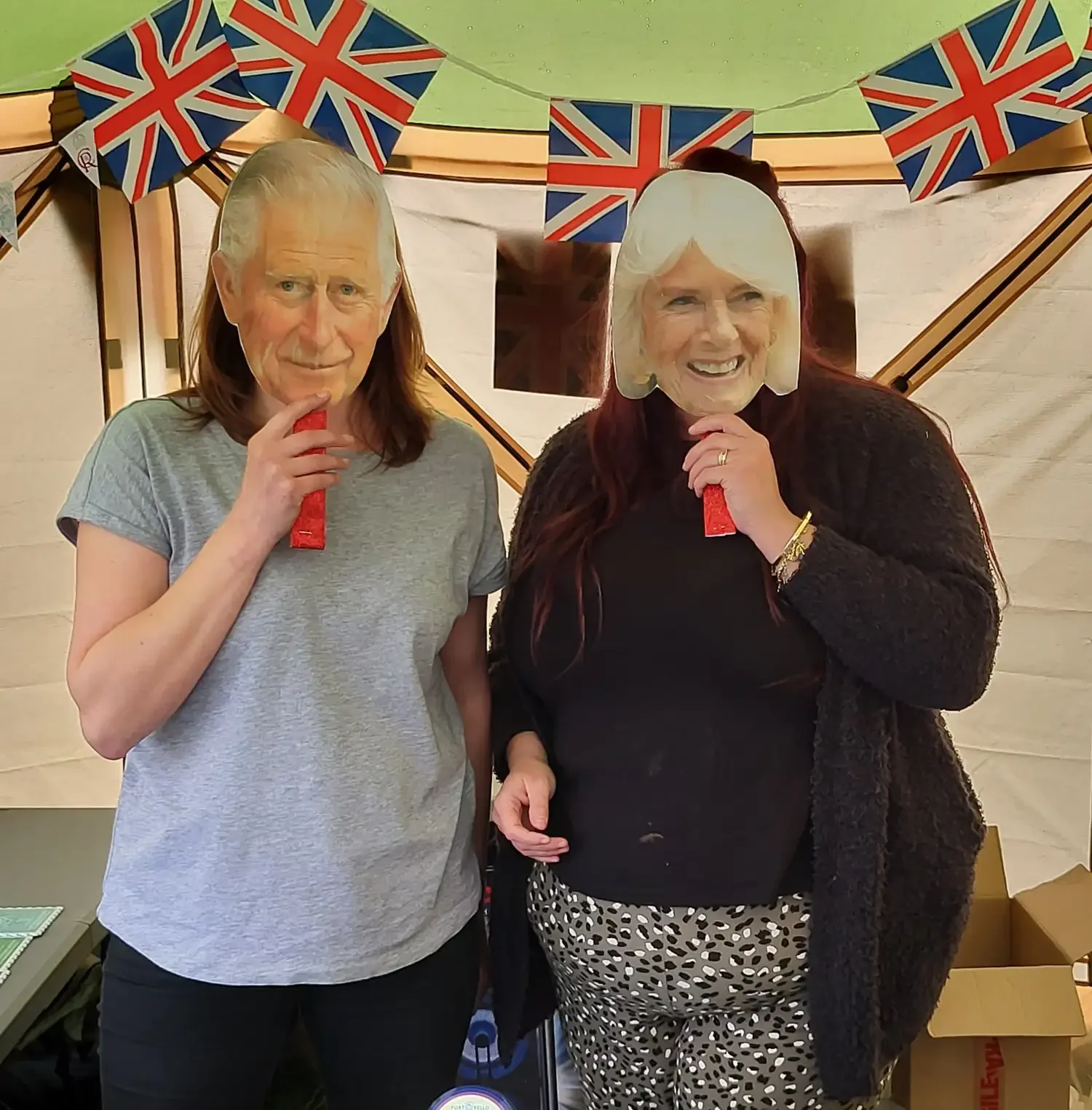 Two women wearing masks with large faces printed on them, celebrating with British flags in the background, inside a tent.