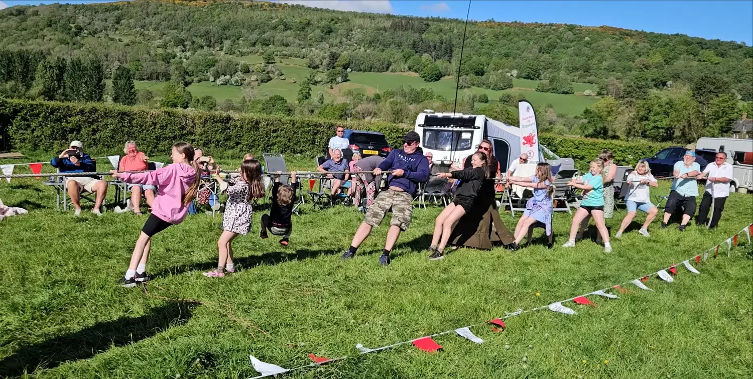 Children and adults participating in a game of tug of war on a grassy field during an outdoor event, with spectators seated and standing in the background, vehicles, and a scenic hilly landscape.