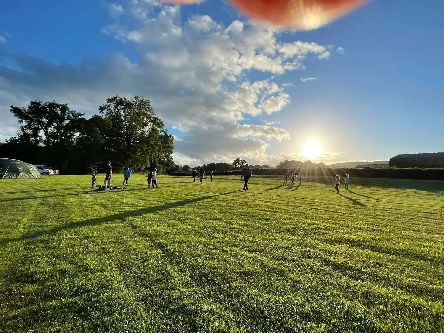 A group of people, including children, playing and walking on a large grassy field during sunset with long shadows, trees in the background, and a partly cloudy sky.