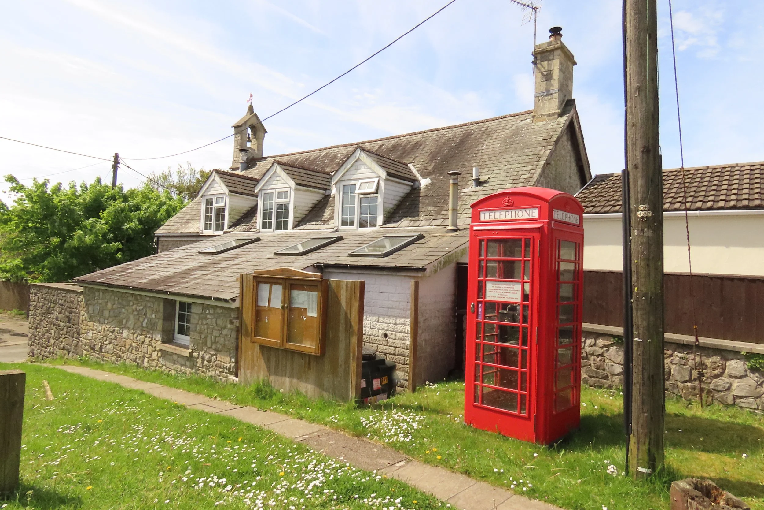 A red British telephone booth in front of a stone and wooden building with multiple skylights and small windows.