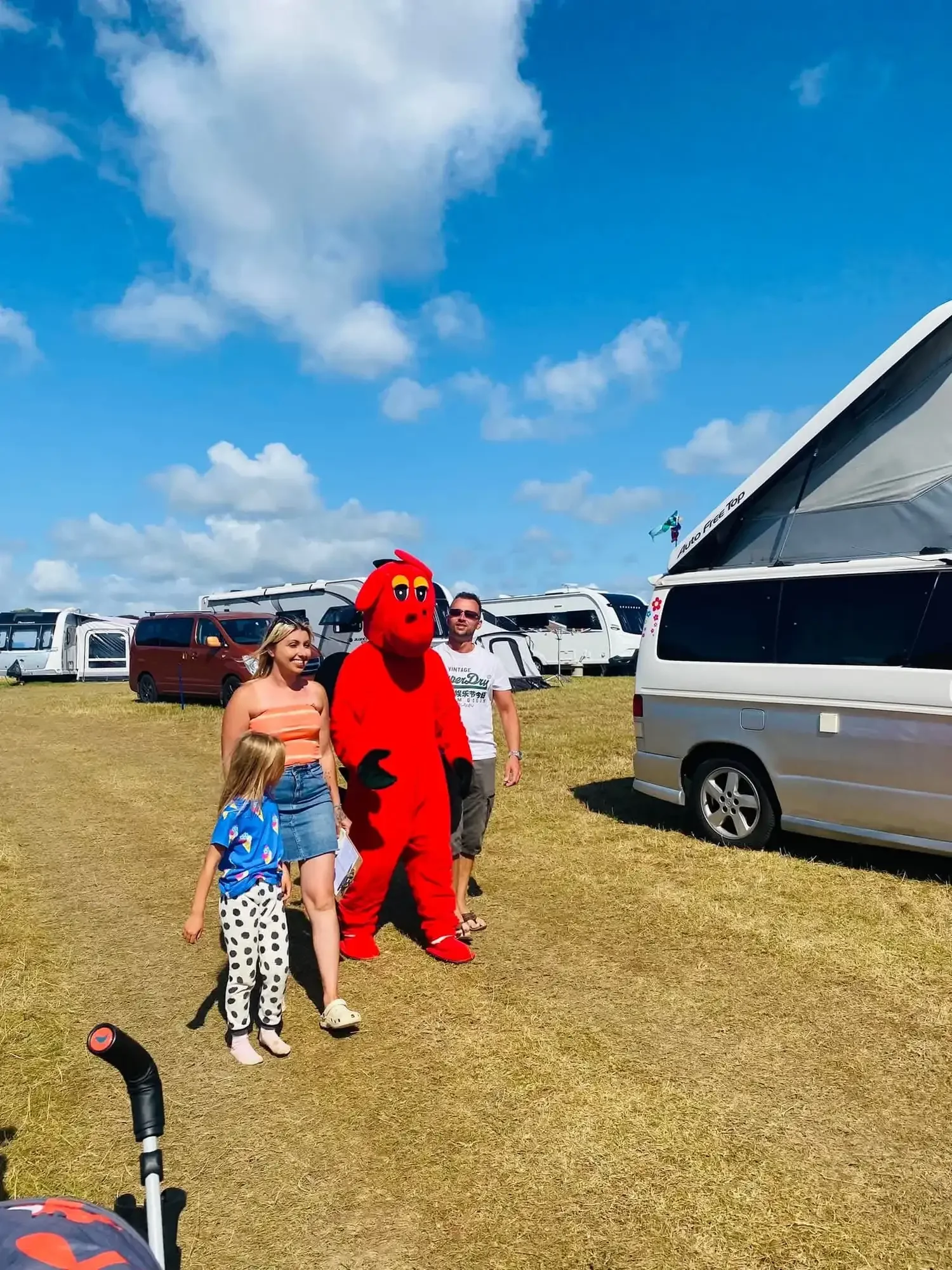 People standing outdoors on a grassy field, including a person in a red mascot costume resembling a cartoonish animal, with balloons and camper vans in the background under a partly cloudy sky.