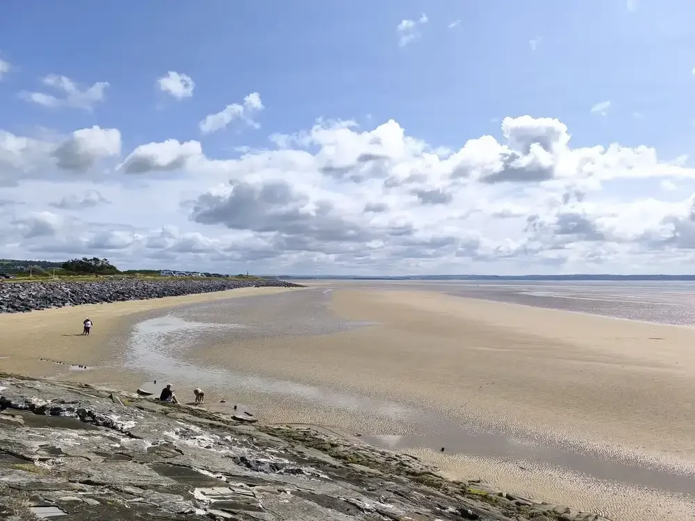 A beach with a wide sandy shoreline under a partly cloudy sky, and a few people sitting and walking on the sand, with a rocky breakwater on the left side of the image.
