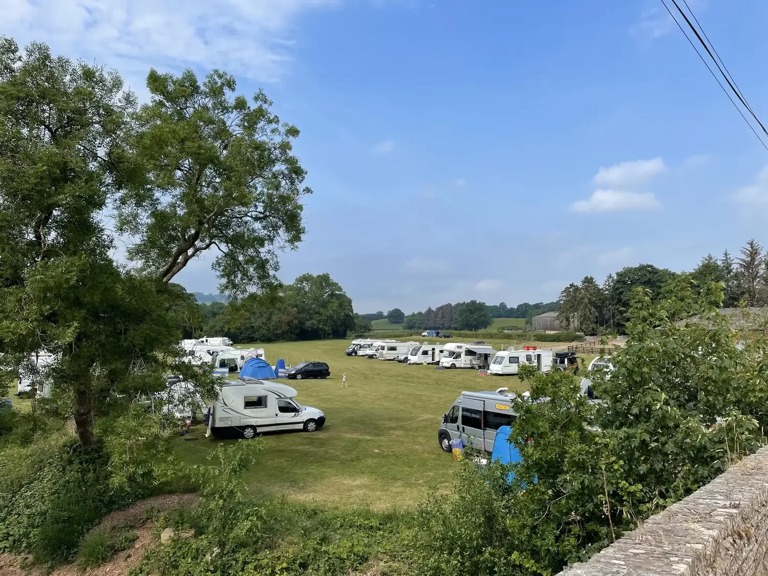 A large grassy field with multiple recreational vehicles (RVs) and tents set up, surrounded by trees under a blue sky with a few clouds.