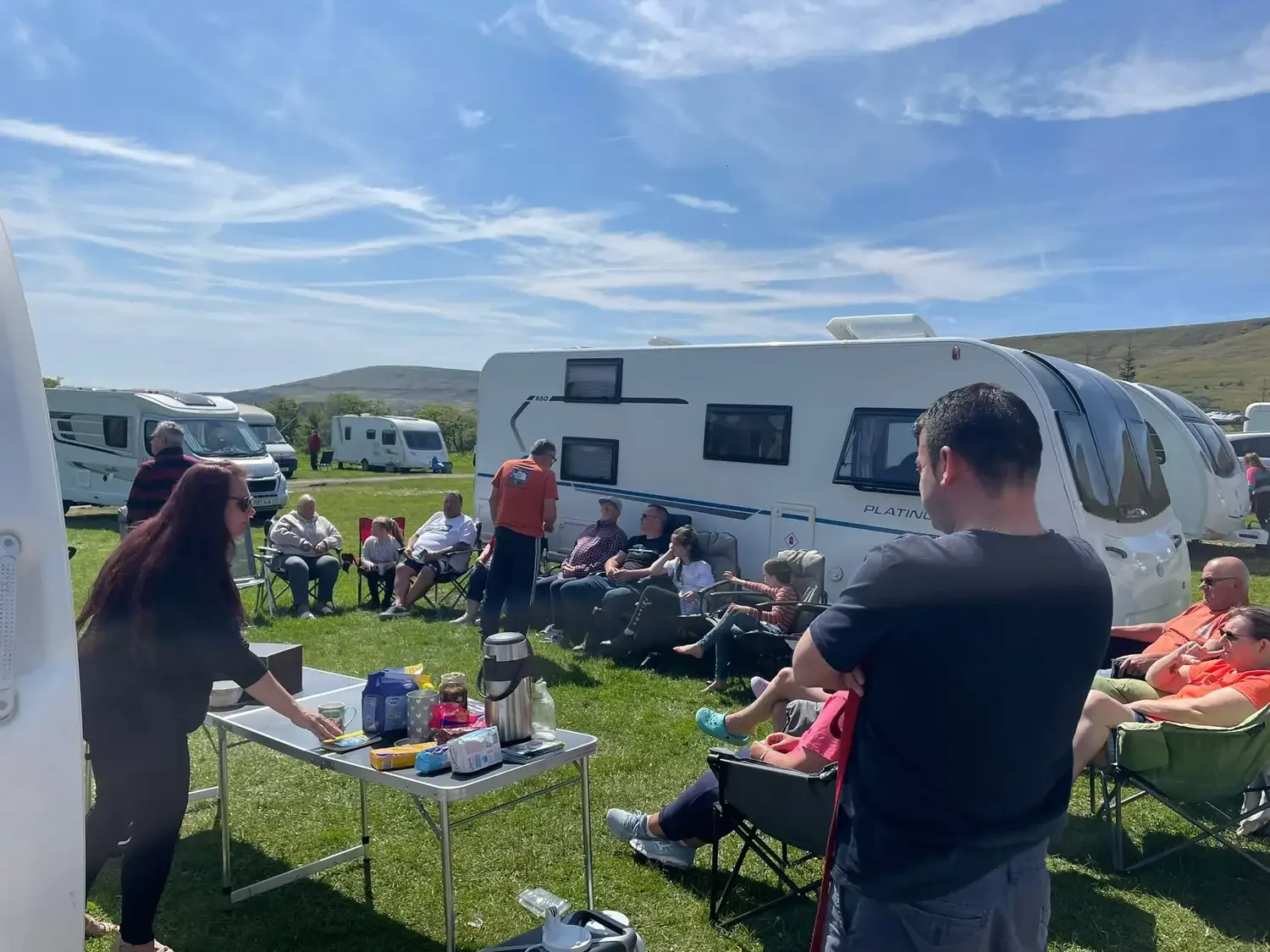 A group of people gathered outdoors at a camping site with caravans, sitting and standing on green grass, enjoying a sunny day with a partly cloudy sky.