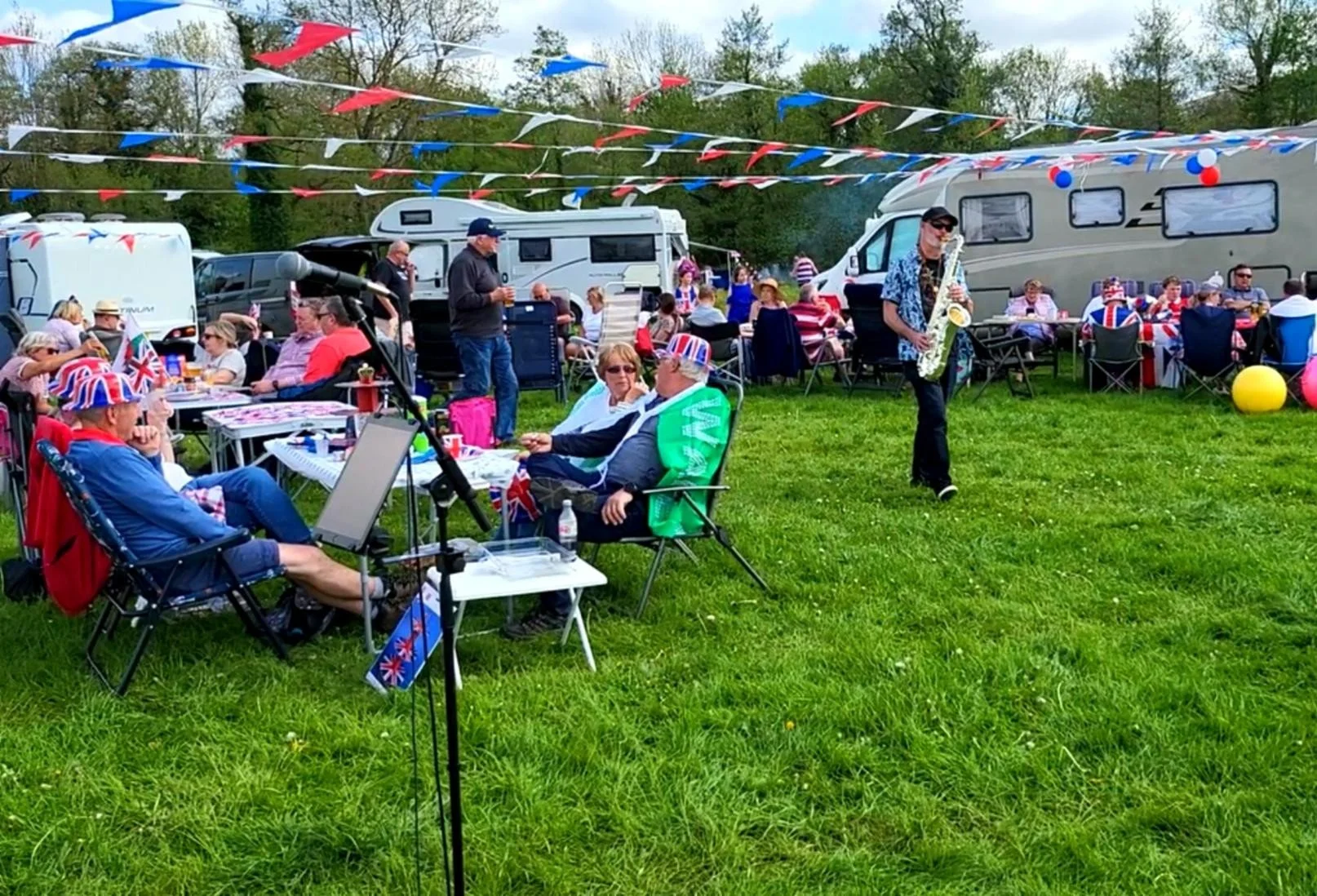 People gathered outdoors celebrating with red, white, and blue decorations, including Union Jack hats and flags, with camping trailers and trees in the background.