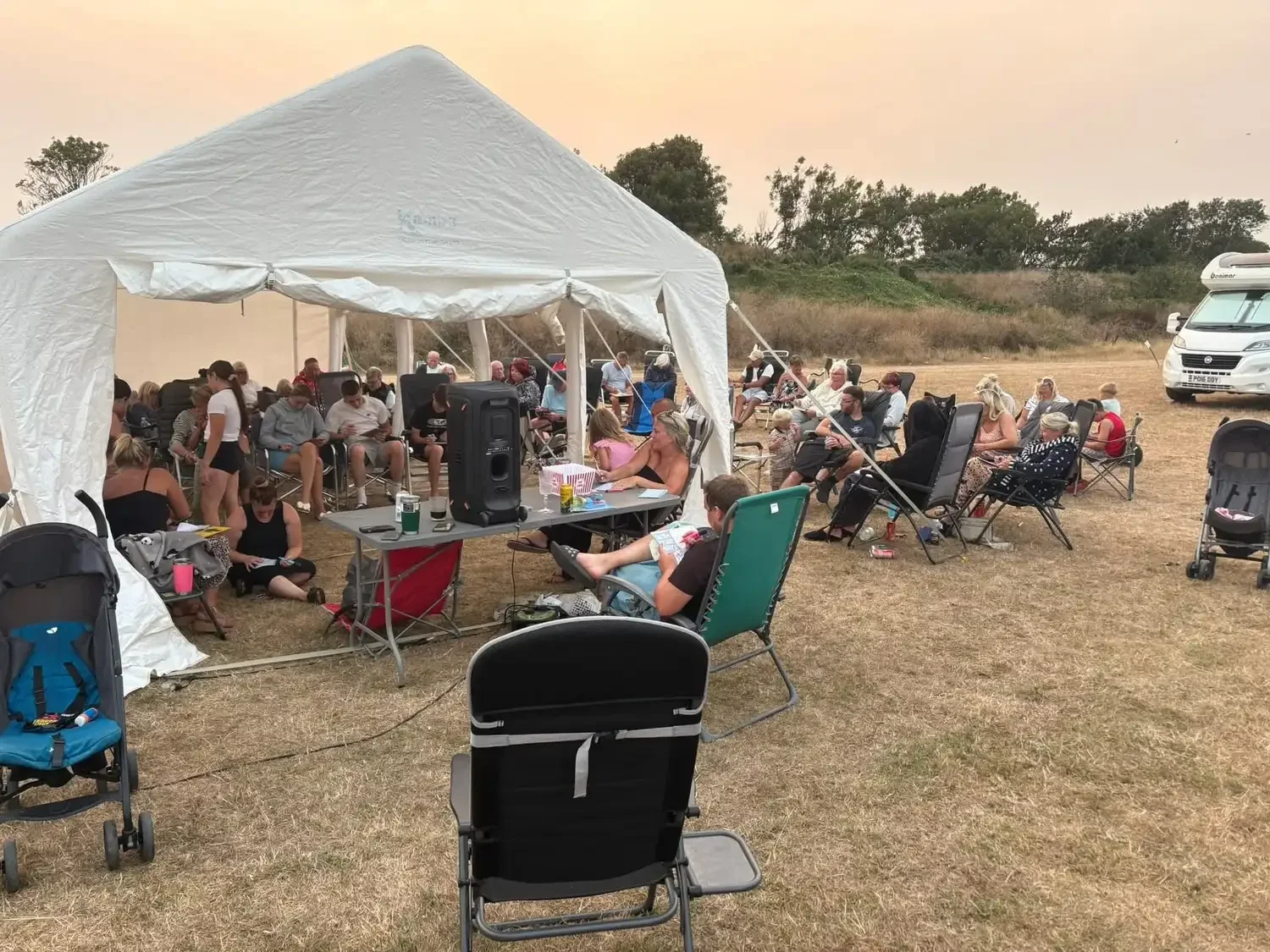 Outdoor gathering under a large white tent with many people sitting in chairs on a dry grassy field, some under the tent and some outside, with a few strollers and a RV in the background.